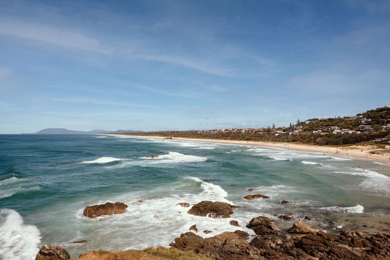 Aerial panoramic view of coastal waves crashing against cliffs along a scenic beach landscape under blue skies.