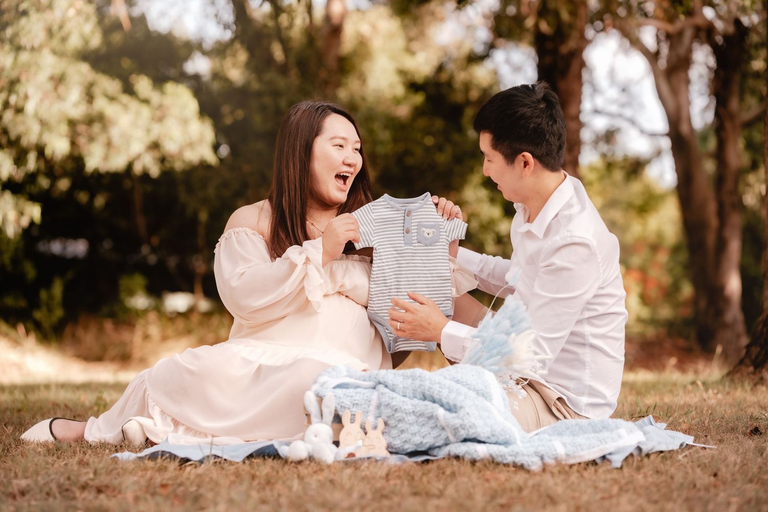 A couple in white outfits sits together on a blanket in a sun-dappled outdoor setting during autumn.