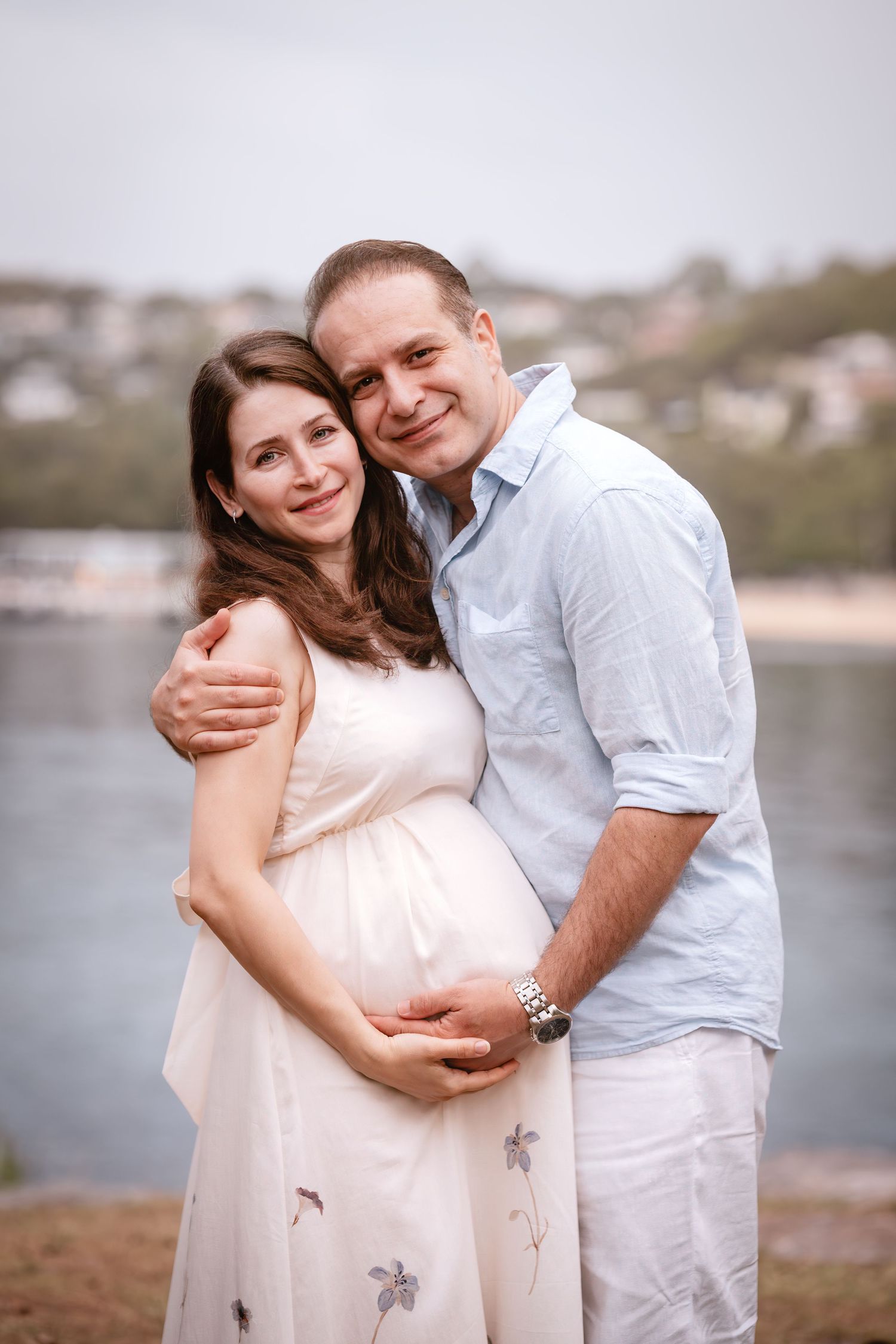 A couple in white outfits embraces by the waterfront during a maternity photoshoot at sunset.
