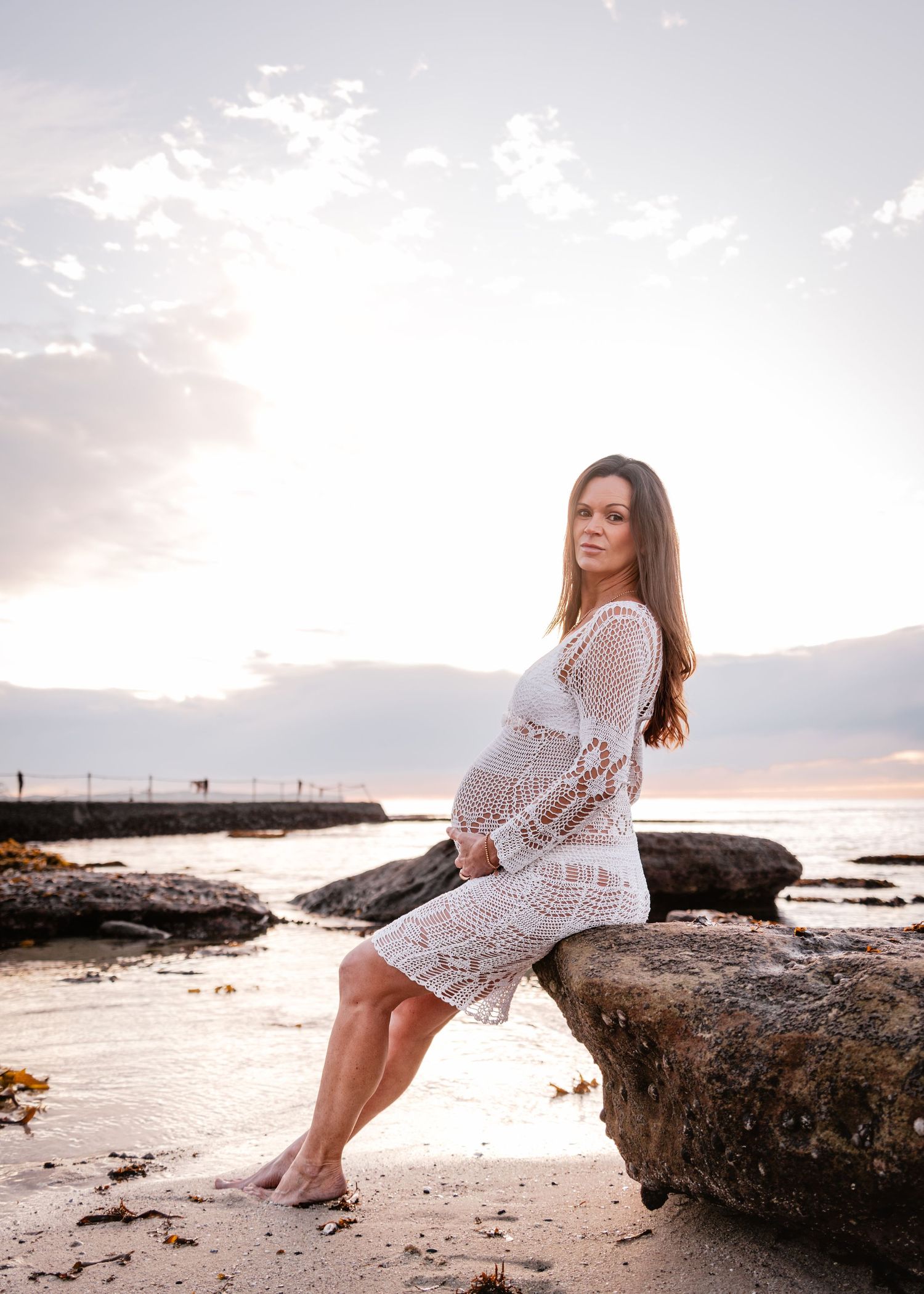 A woman in a white crochet dress poses on rocky coastal formations at sunset by the ocean.