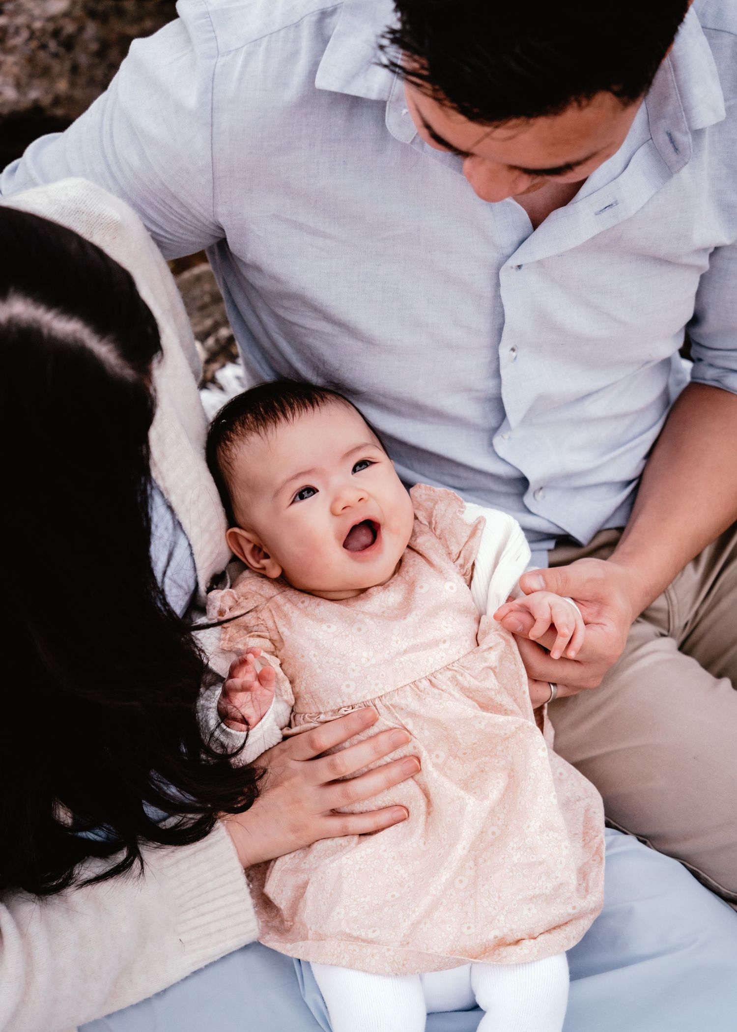 A baby in a peach-colored dress sits on a couch while being held by someone in a light blue shirt.