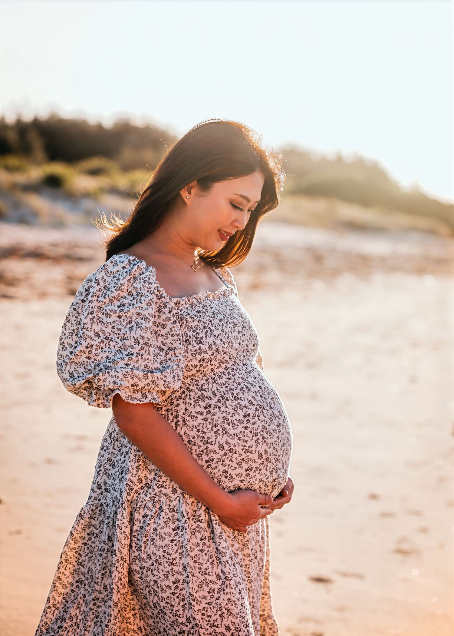 A pregnant woman in a floral dress poses on a beach at sunset with warm golden light illuminating the scene.