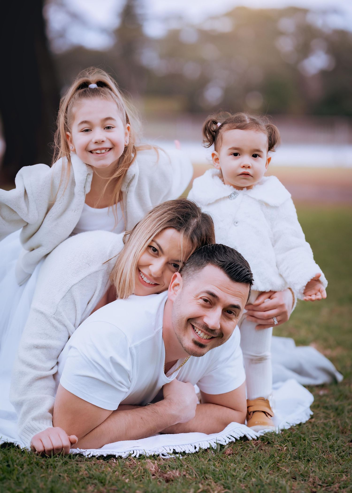Family wearing white outfits smiles while laying together on grass during outdoor portrait session.