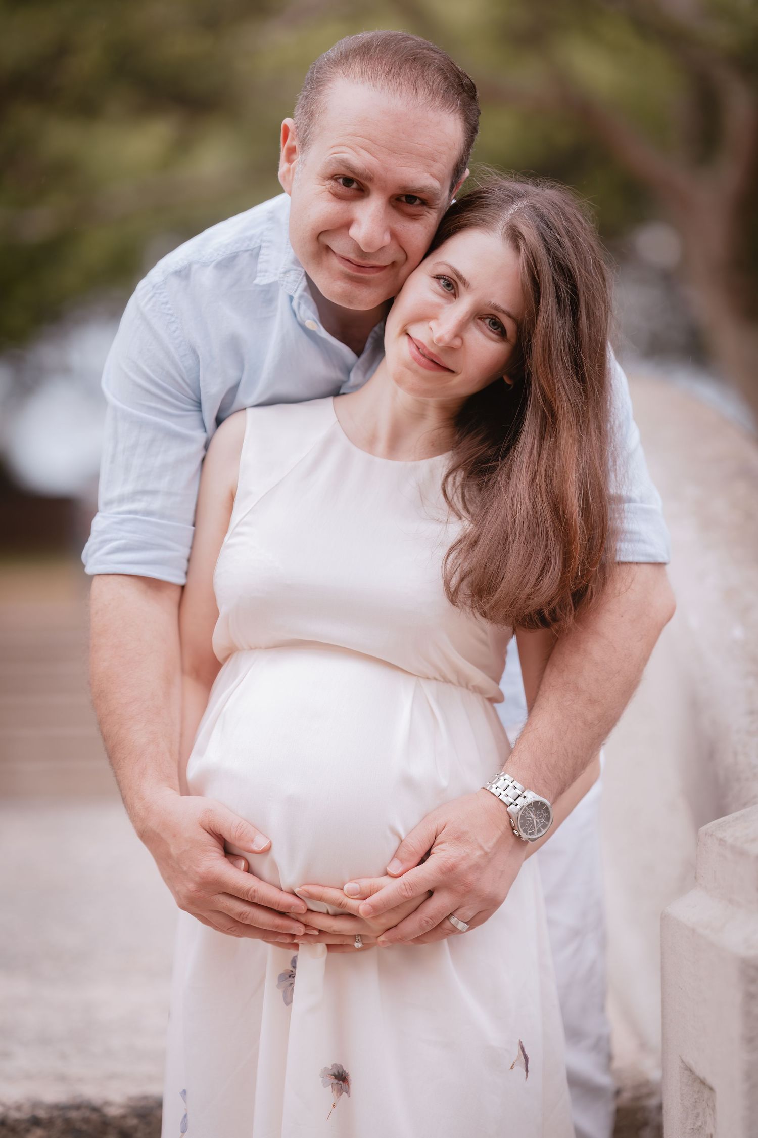 A couple embraces during a maternity photoshoot outdoors, wearing white and grey casual attire.