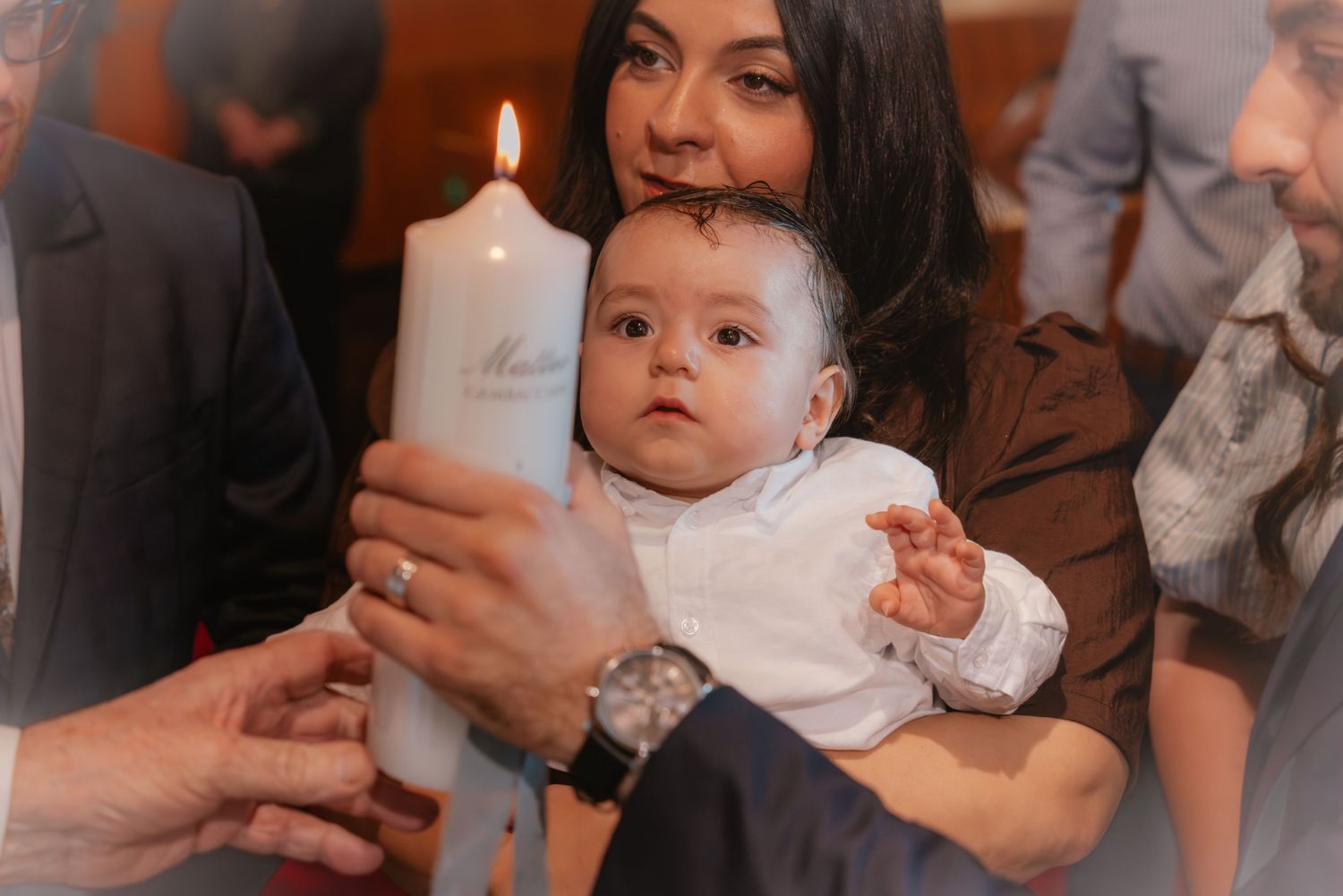 A baby in white baptism outfit holds a christening candle with a woman during a religious ceremony.
