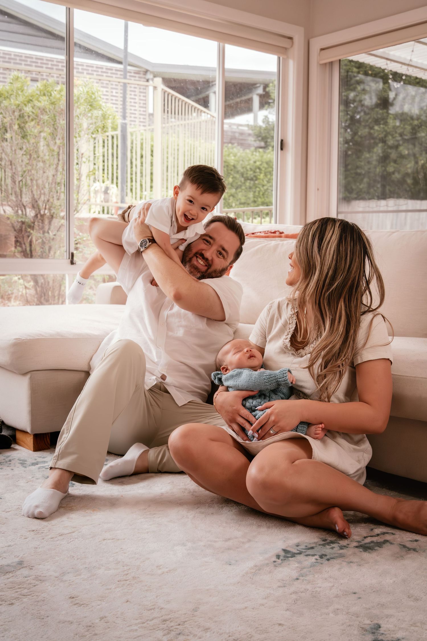 Happy family sitting on living room floor, dad holding toddler boy while mom cradles newborn baby in bright home.