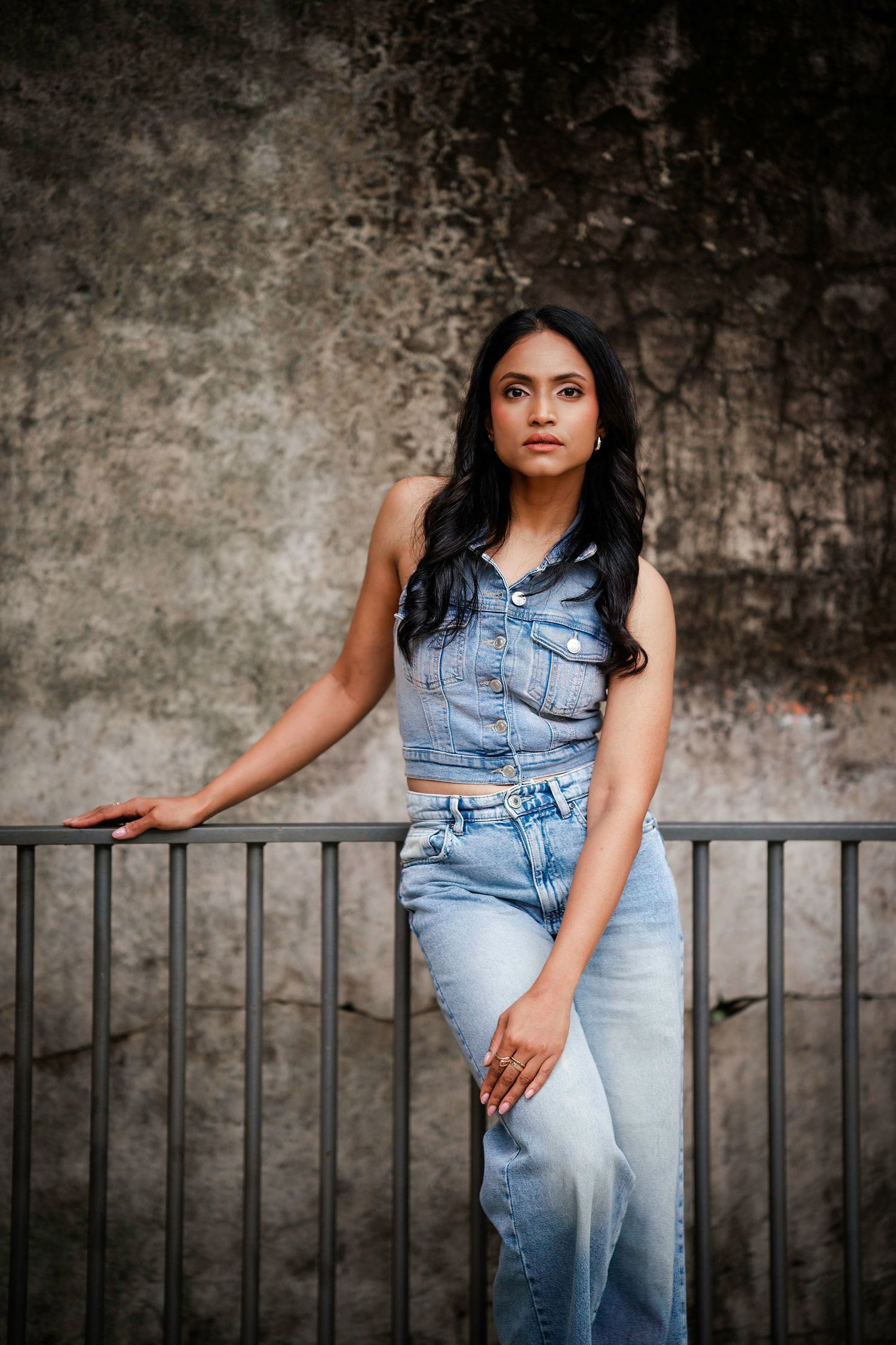 Woman in denim crop top and jeans poses against textured concrete wall with metal railing.