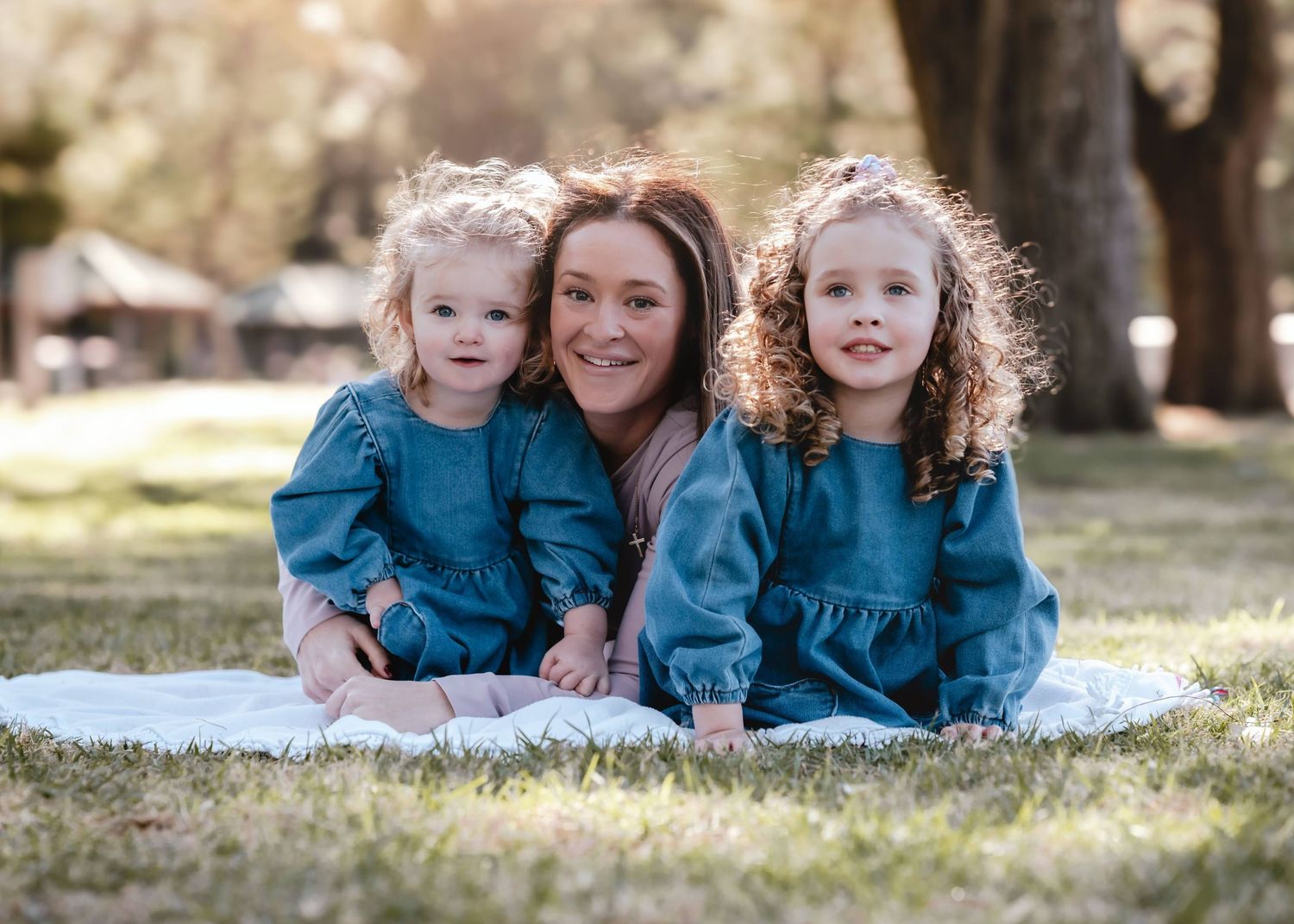 Family wearing coordinating teal dresses sits together on white blanket in sunny outdoor park setting.