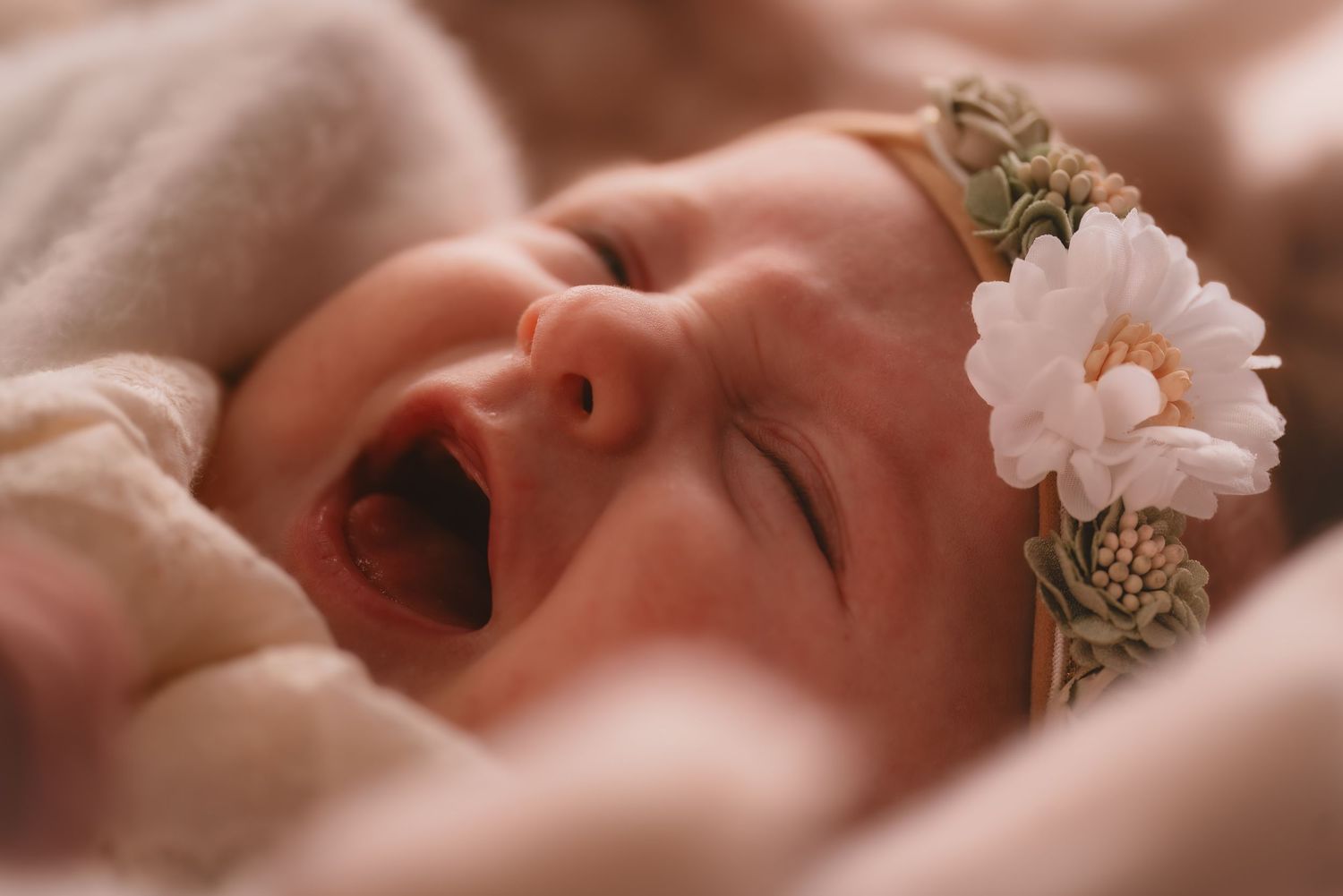 A yawning baby wearing a delicate white flower headband rests on soft pink blankets.