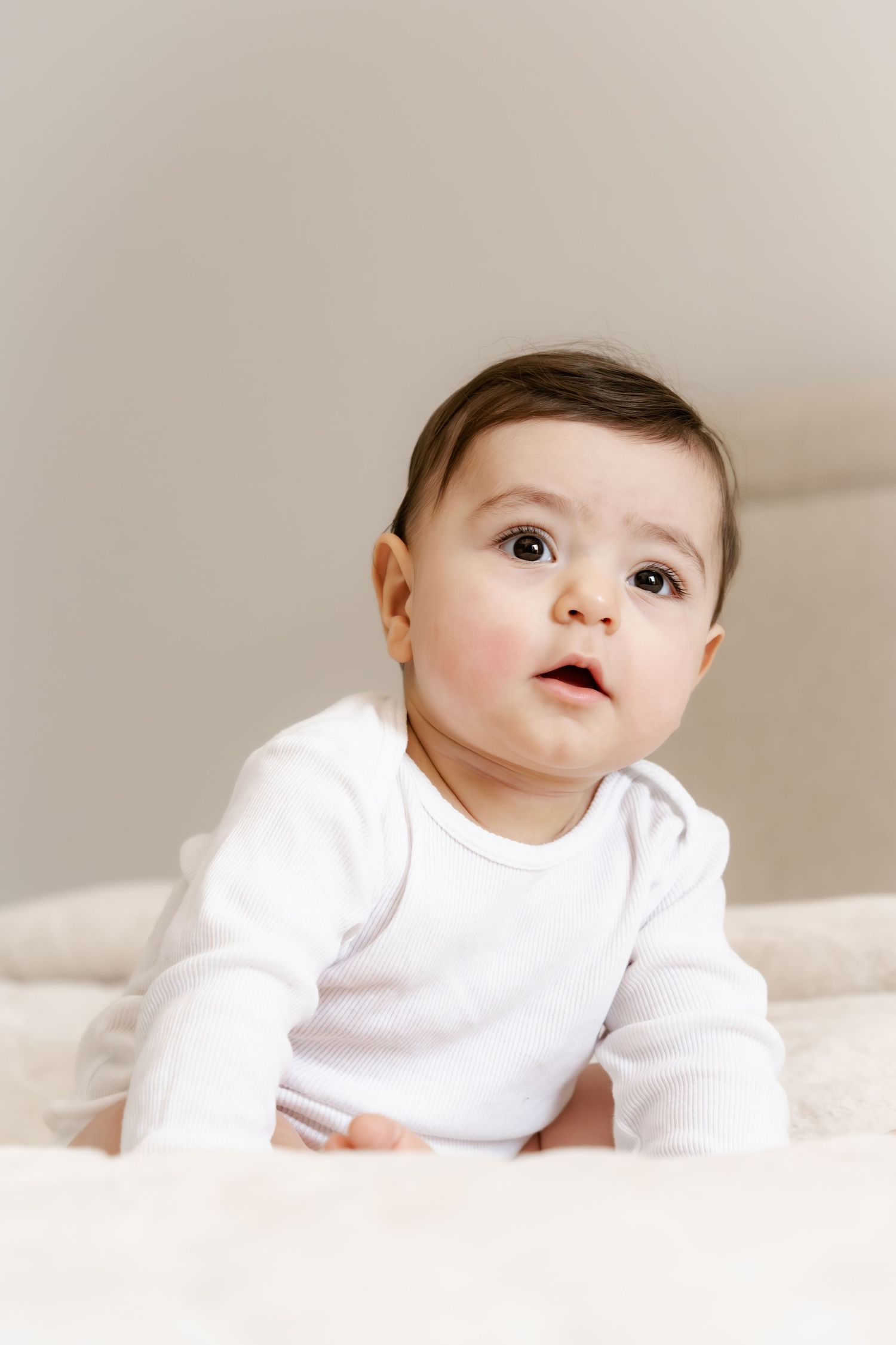 Adorable baby in white onesie sitting up and looking curiously upward with big brown eyes.