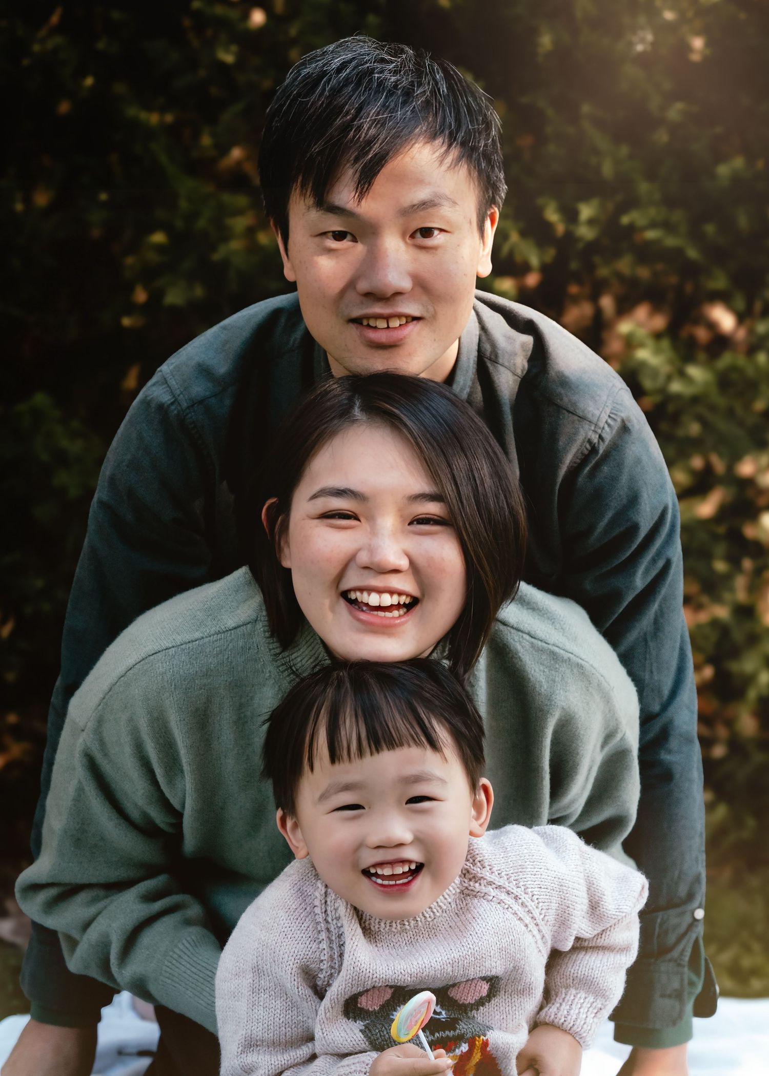A family of three wearing cozy sweaters poses together outdoors with bright genuine smiles in natural lighting.