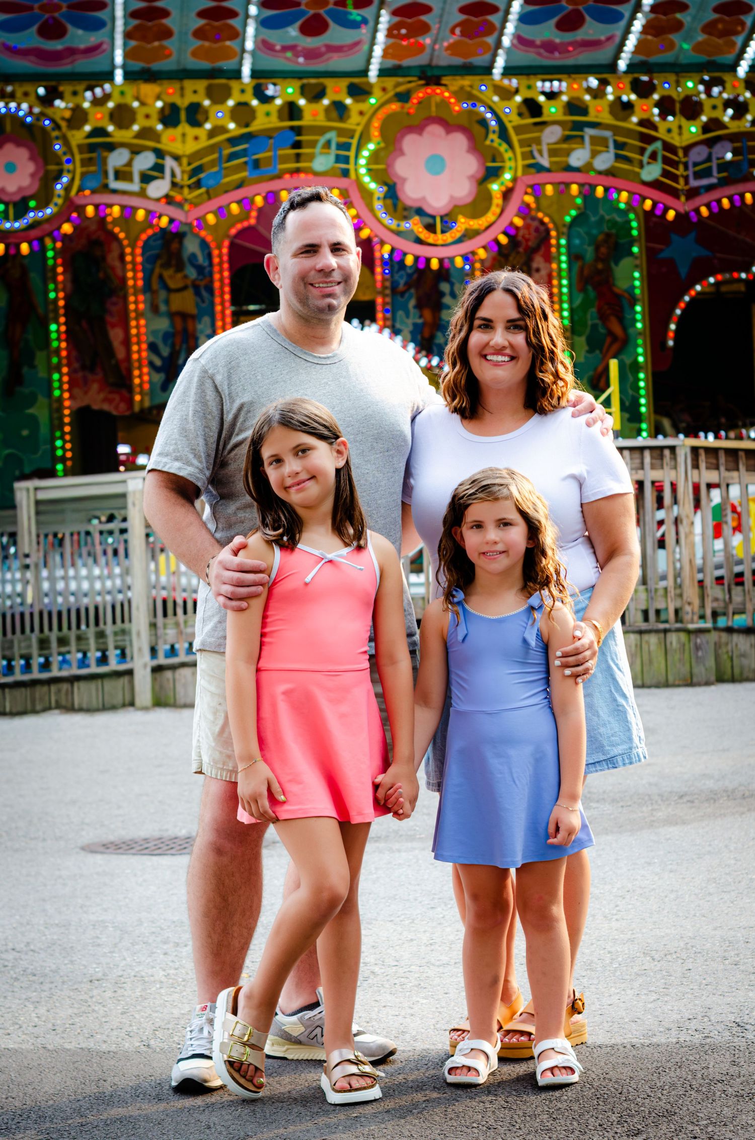 Family of four smiling in front of a colorful carousel at an amusement park on a sunny day.