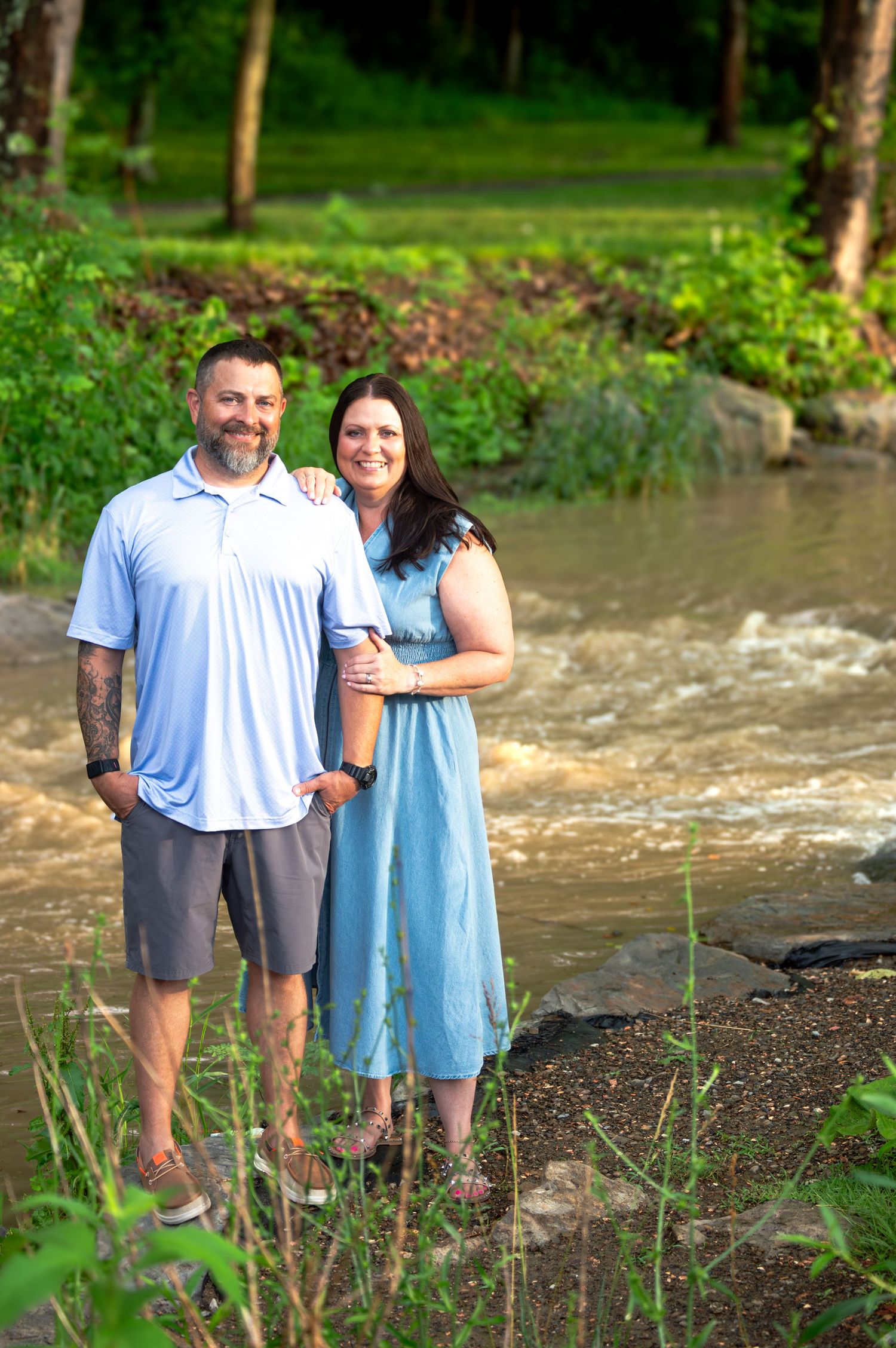 Two people stand together near a creek surrounded by trees and greenery.