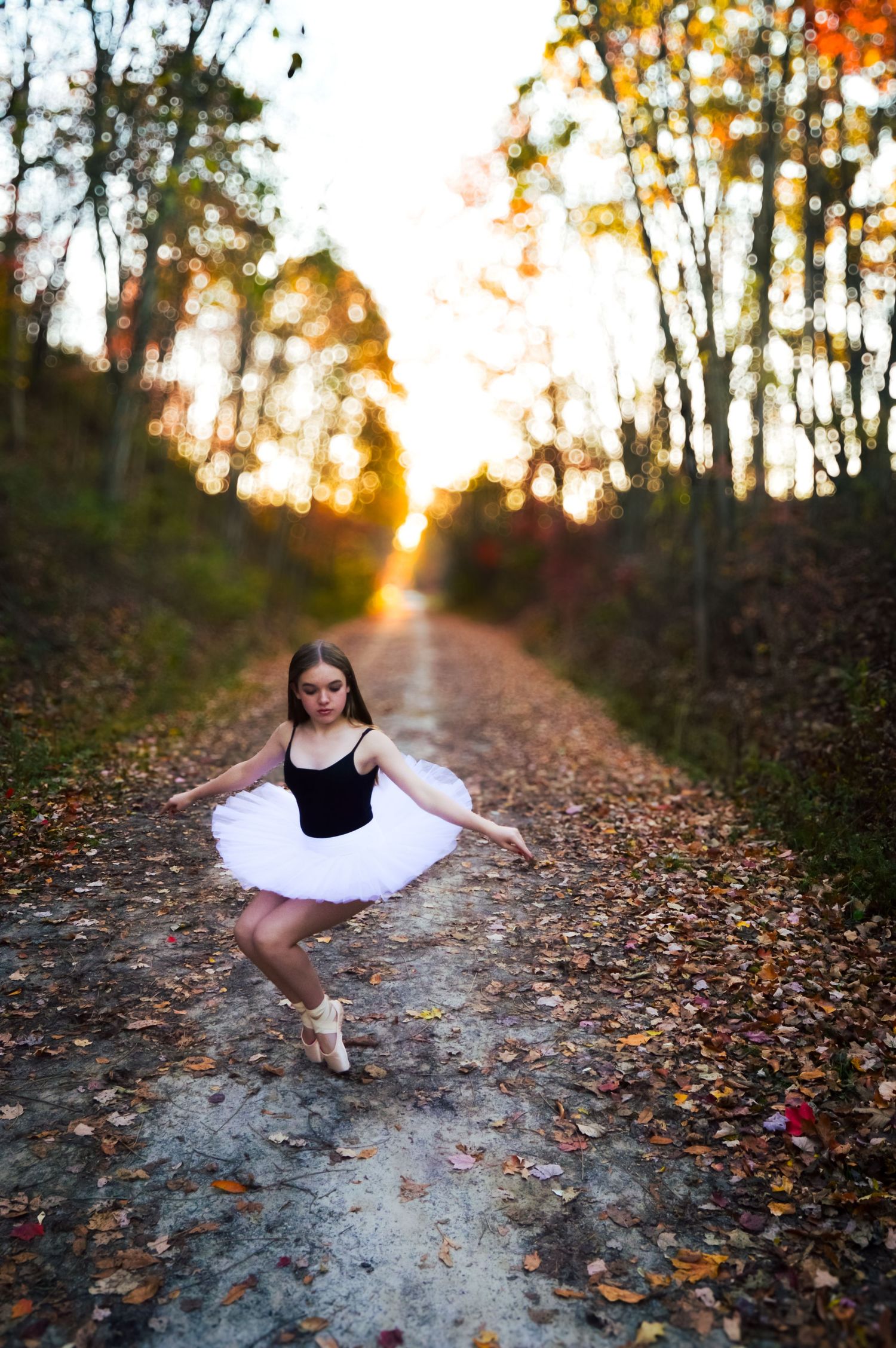 Dancer in white tutu performs on leaf-covered path during autumn sunset.