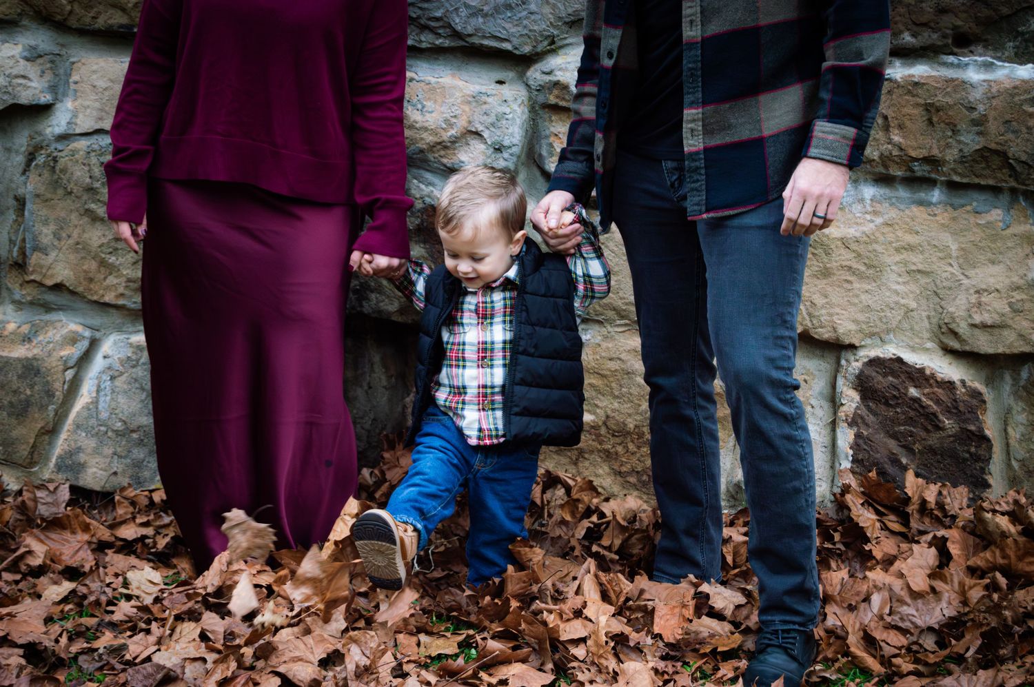 A group of people stand together in autumn leaves with a stone wall backdrop during an outdoor family photoshoot.