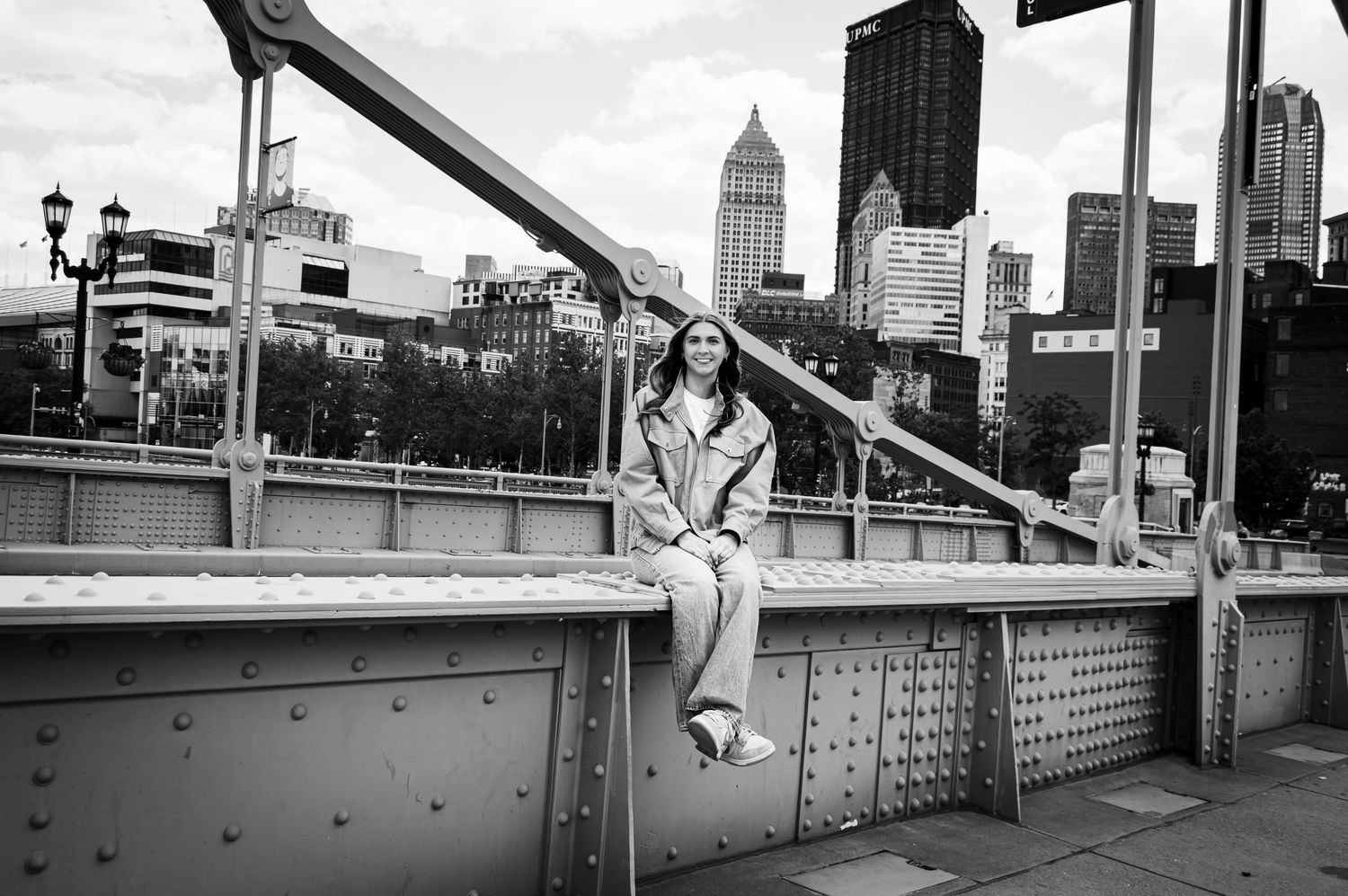 Black and white photo of woman sitting on bridge with city skyline in background.