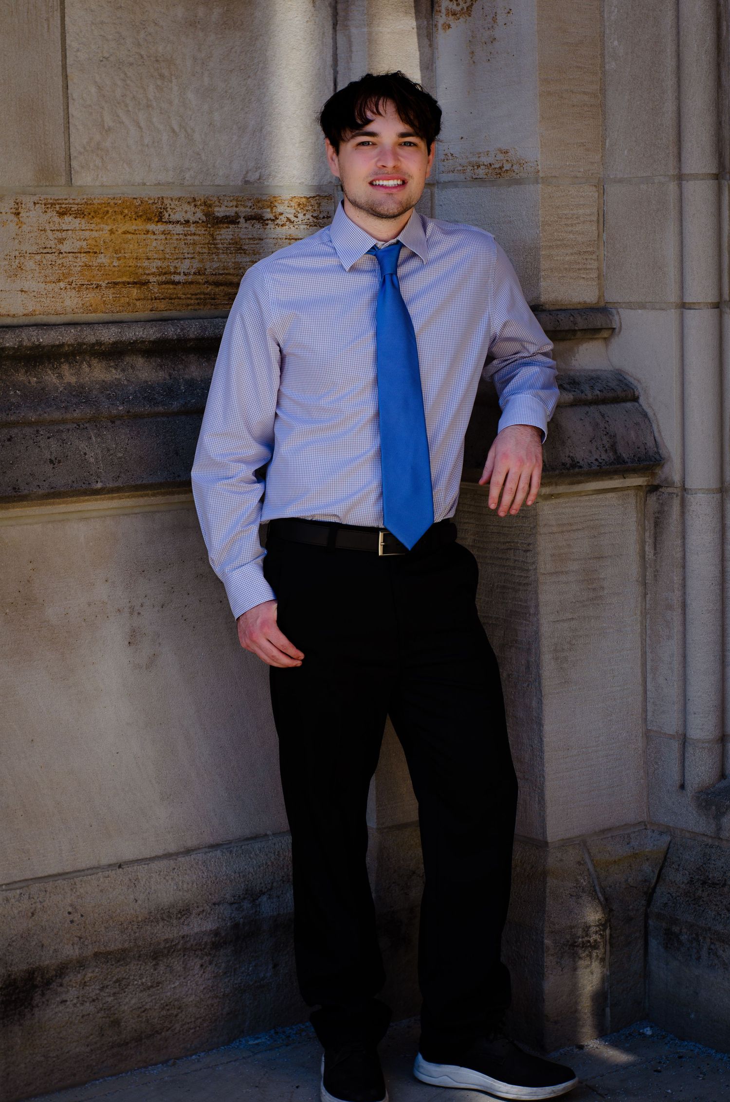 Young man in blue tie and dress shirt leans against stone column in professional portrait photo.