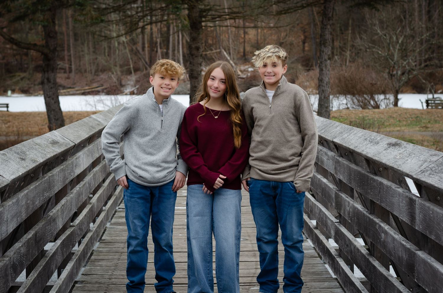 Three people in casual clothing sit together and laugh while posing on a wooden bridge in a winter setting.