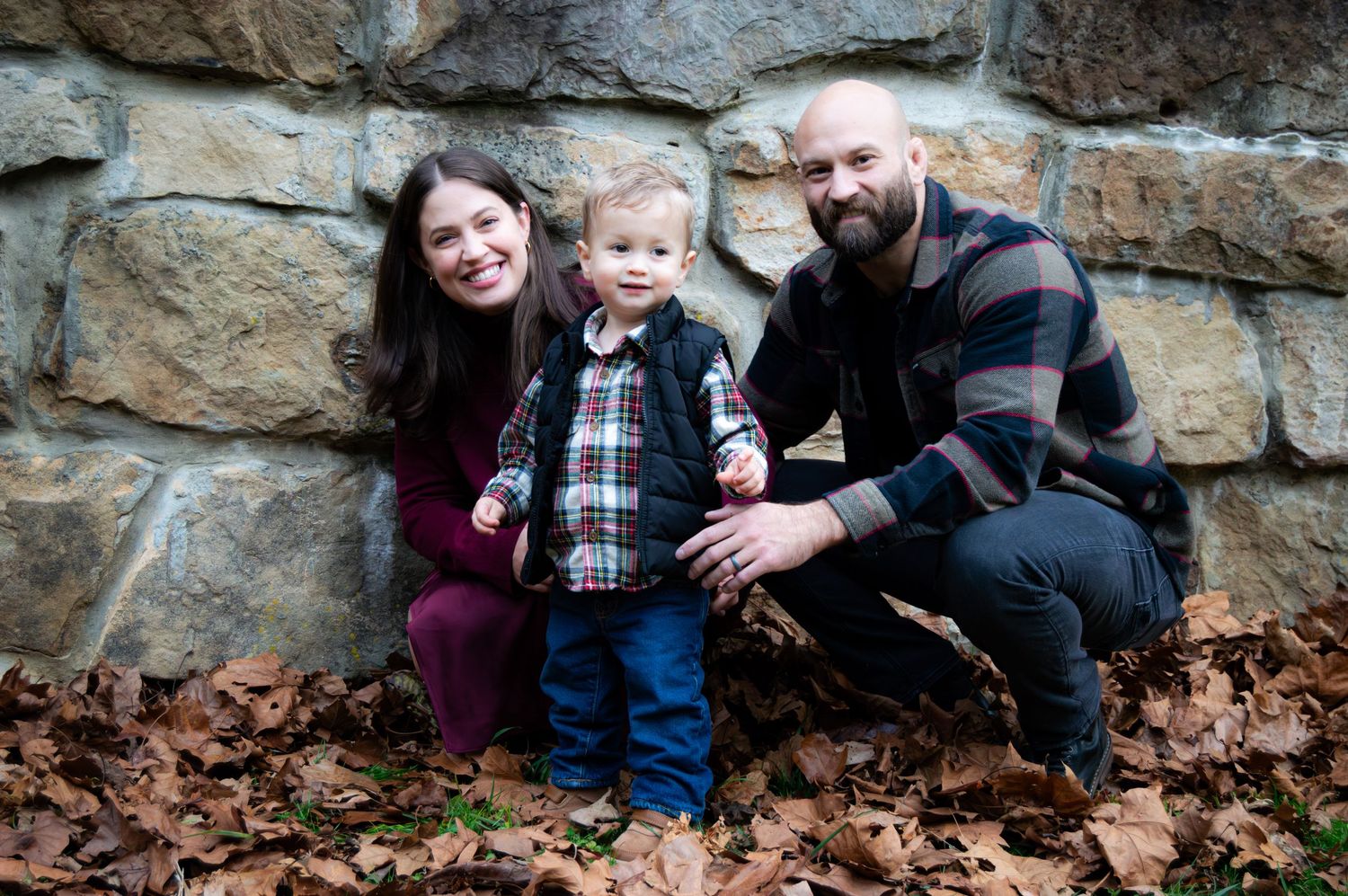 Family embraces while sitting together on fallen leaves by a rustic stone wall.