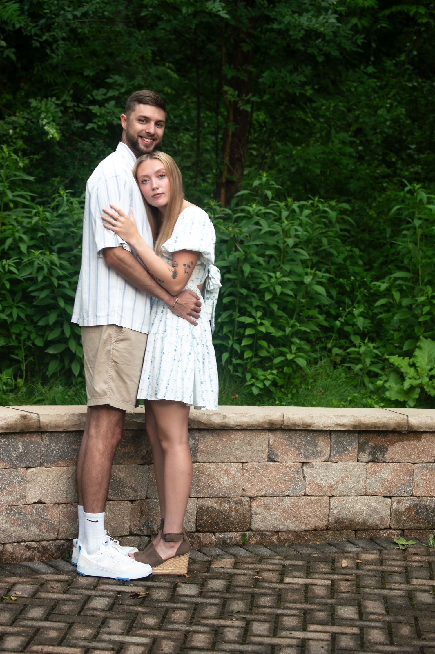 Couple in casual summer attire posing against a stone wall with green foliage background.