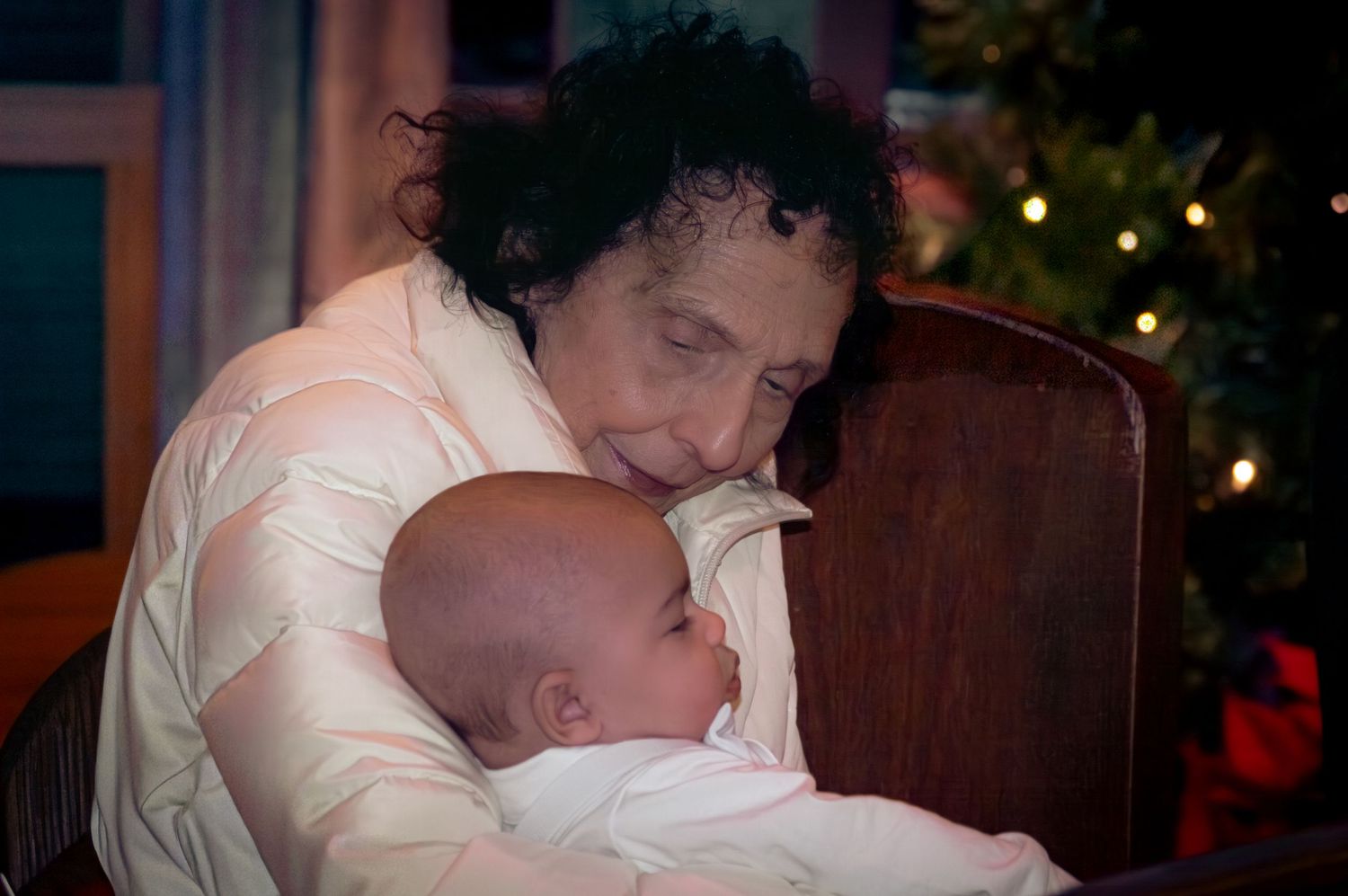 A tender moment between a caregiver and sleeping baby with Christmas lights glowing in the background.