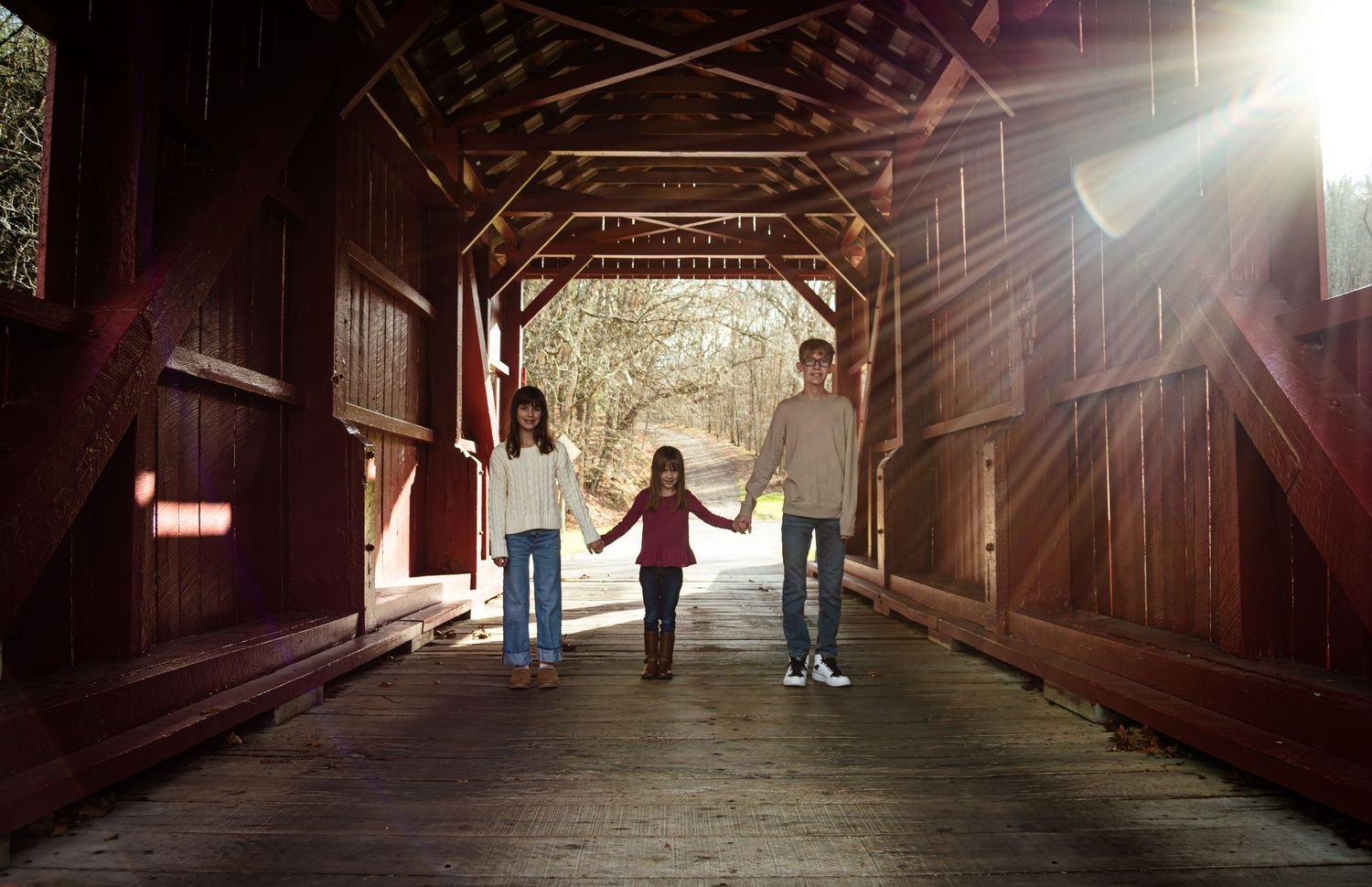 Three people hold hands while walking through a sunlit wooden covered bridge with beams of light streaming in.
