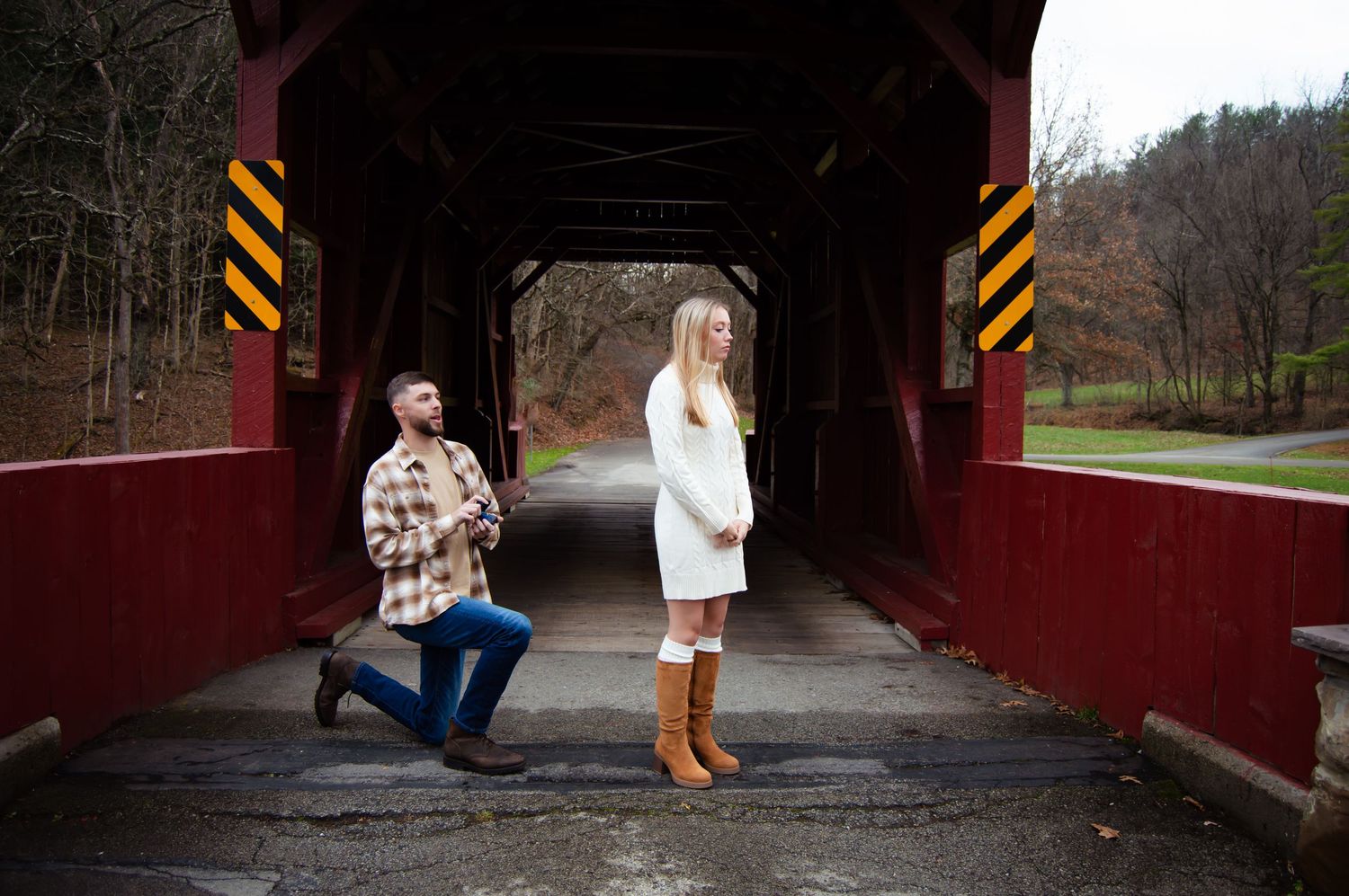 A couple poses for engagement photos at a rustic red covered bridge in autumn.