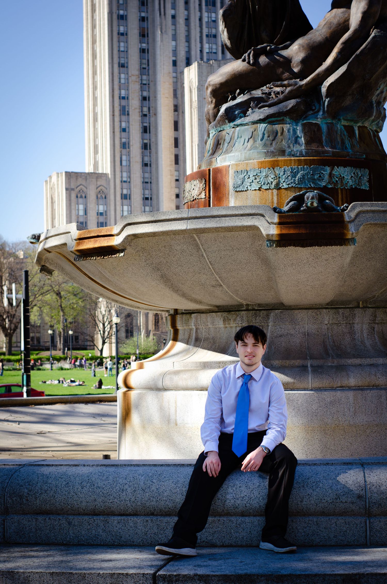 Young man in white shirt and blue tie sits on ornate fountain base with tall building and park in background.