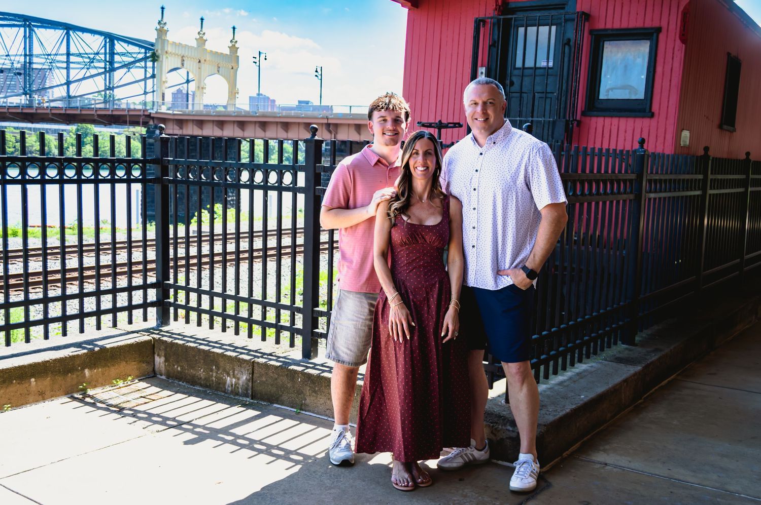 Two men and a woman pose together on an outdoor deck near a red building and iron fence.