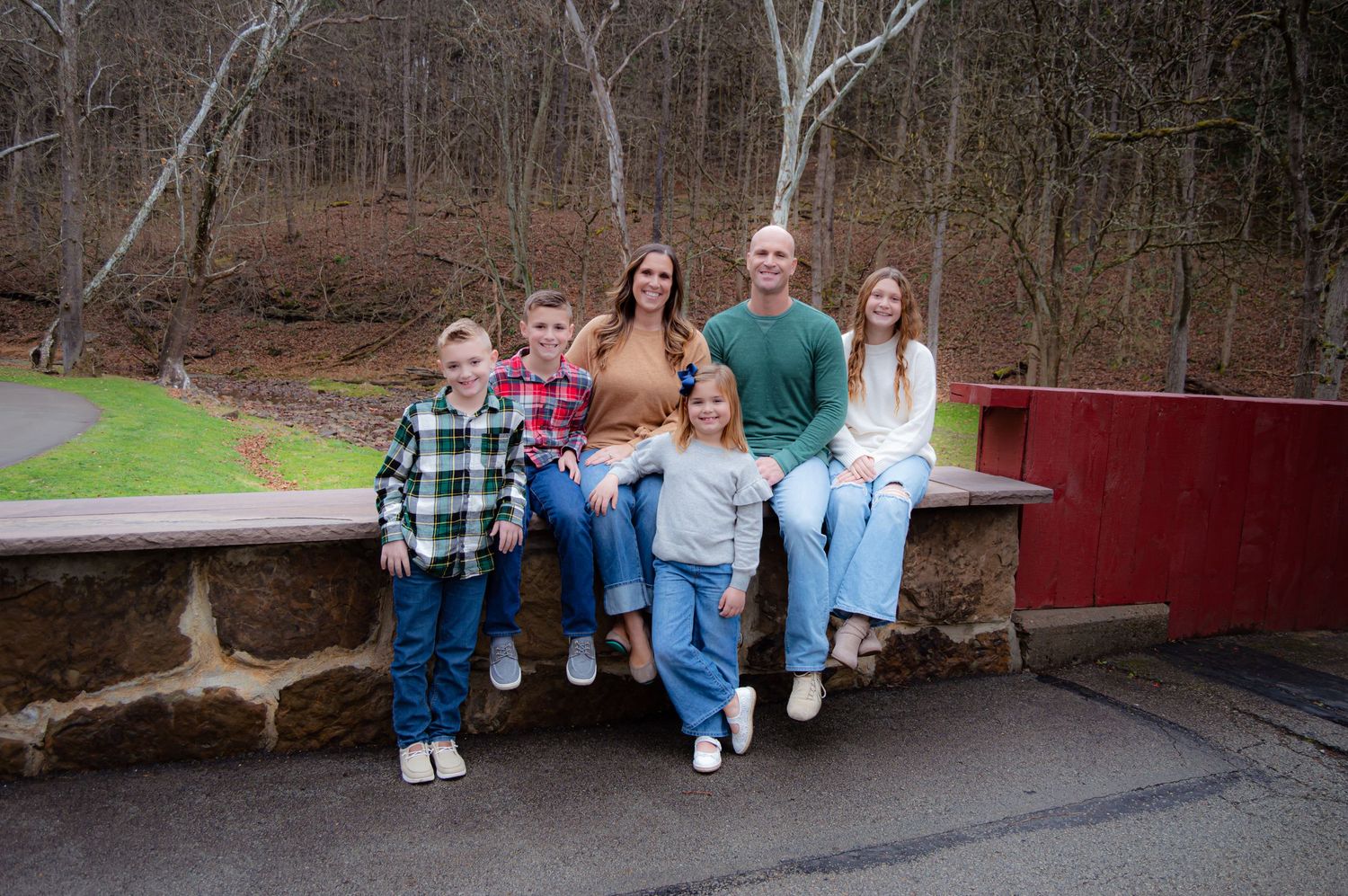 A group of people sitting on a wooden fence in a wooded outdoor setting during autumn.