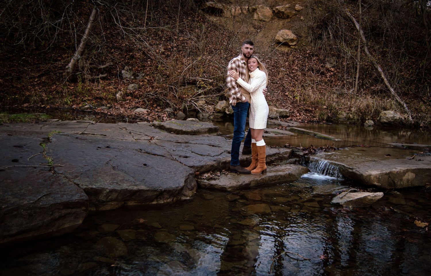 A couple stands at the edge of a creek in a wooded area, enjoying a peaceful moment together in nature.