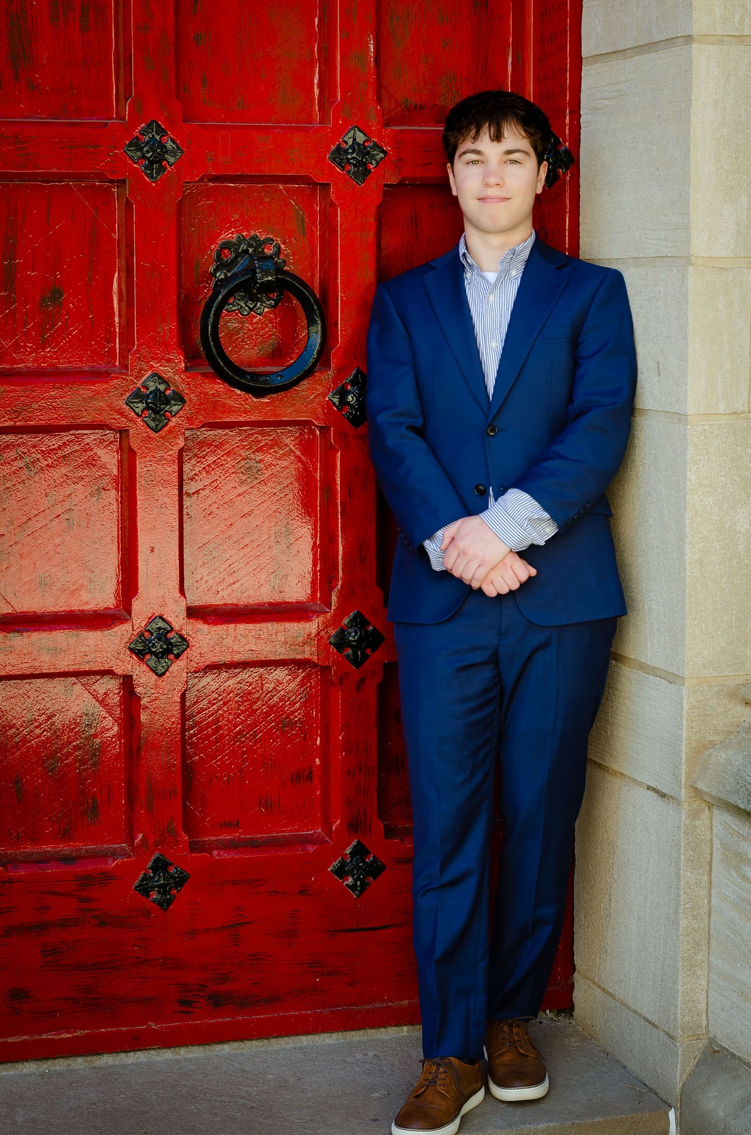 Young man in blue suit posing beside a rustic red door with iron hardware against a stone wall.