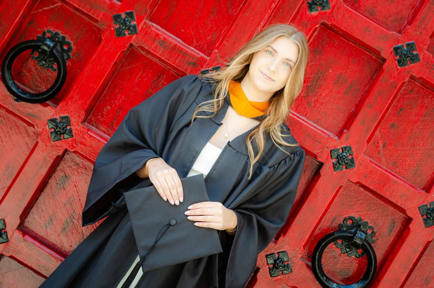 Blonde woman in black academic graduation gown with orange hood holds cap in front of red ornate doors.