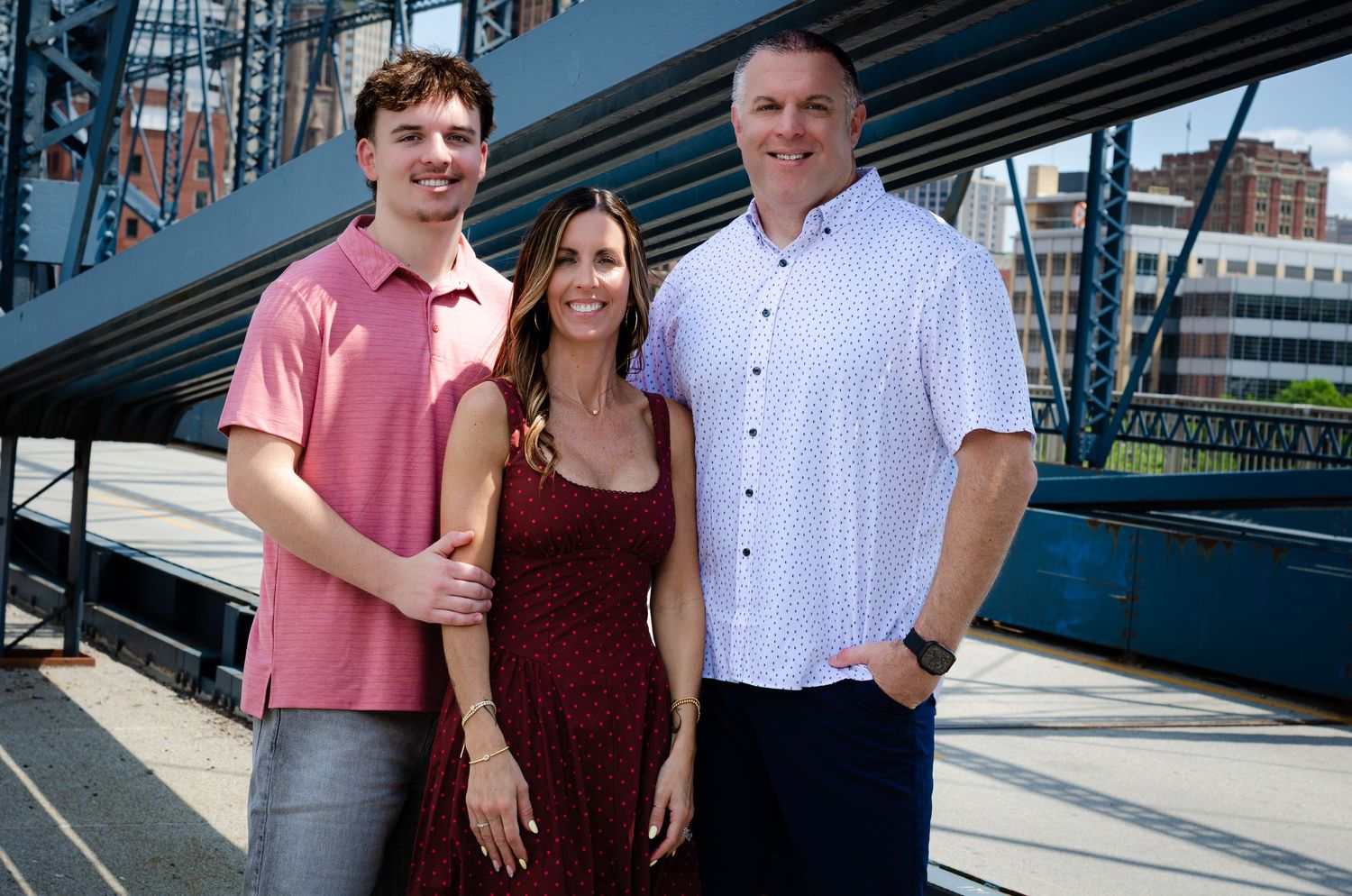 Family of three smiling together on a bridge with city skyline in the background.