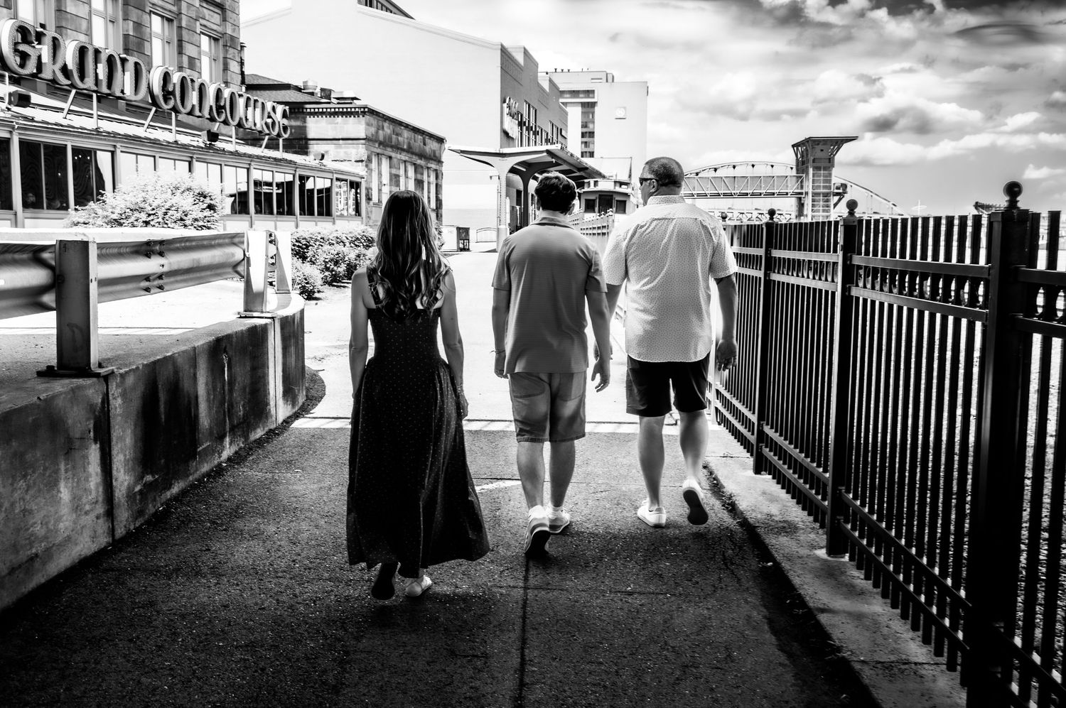 Black and white photo of three people walking along a boardwalk sidewalk past a railing and buildings.