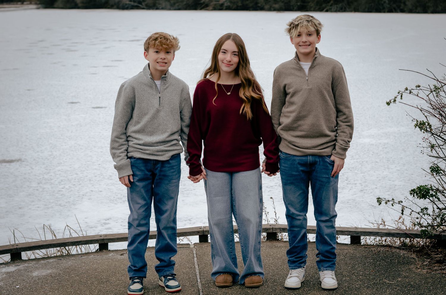 Three siblings in matching sweaters and jeans stand together for a casual outdoor family photo session.