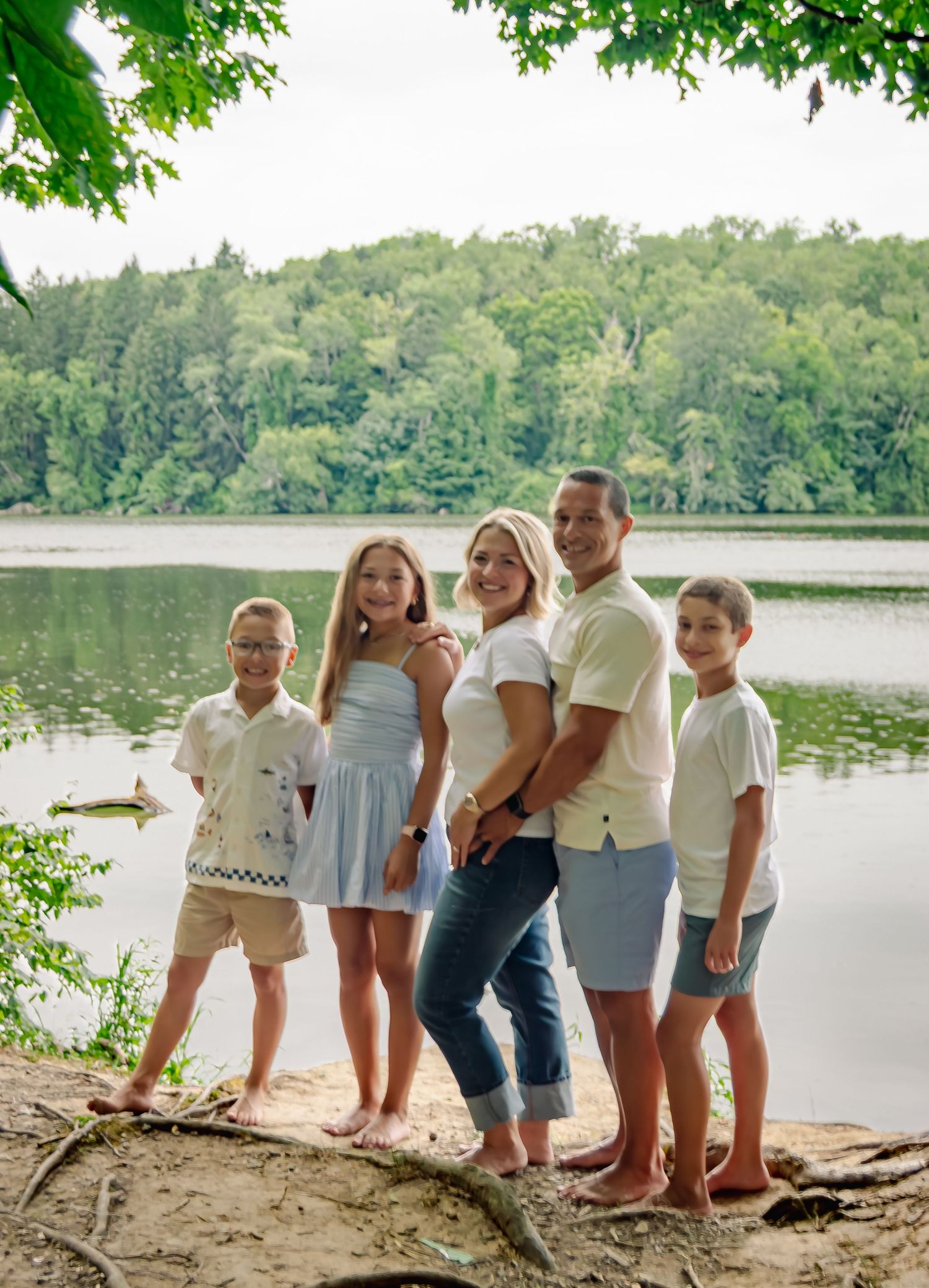 Casual outdoor family gathering on a sandy beach beside a peaceful lake with green foliage overhead.