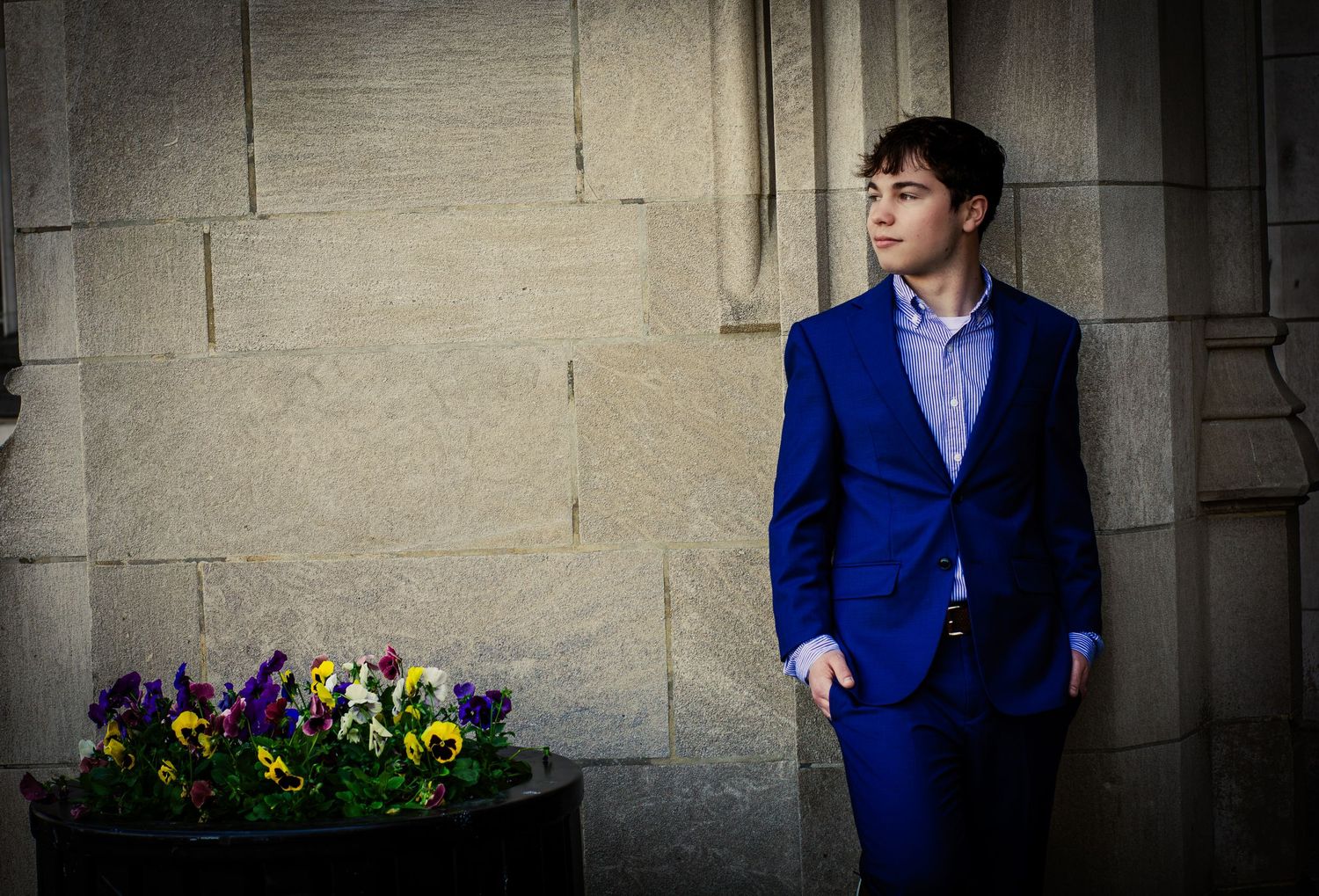 Young man in a royal blue suit leaning against a stone wall with colorful pansy flowers nearby.