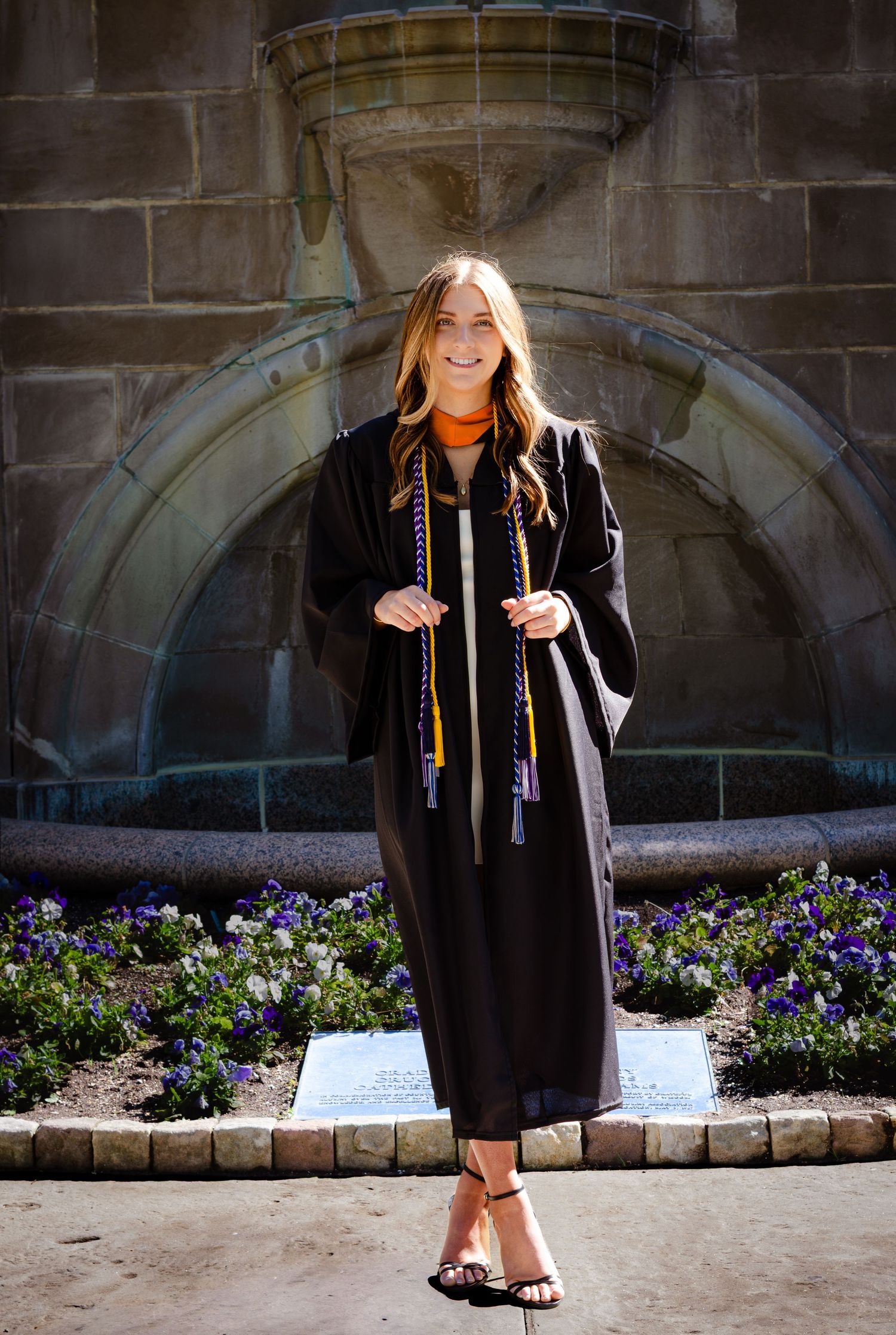 Young woman smiling in black graduation gown with honor cords standing before stone fountain with purple flowers.