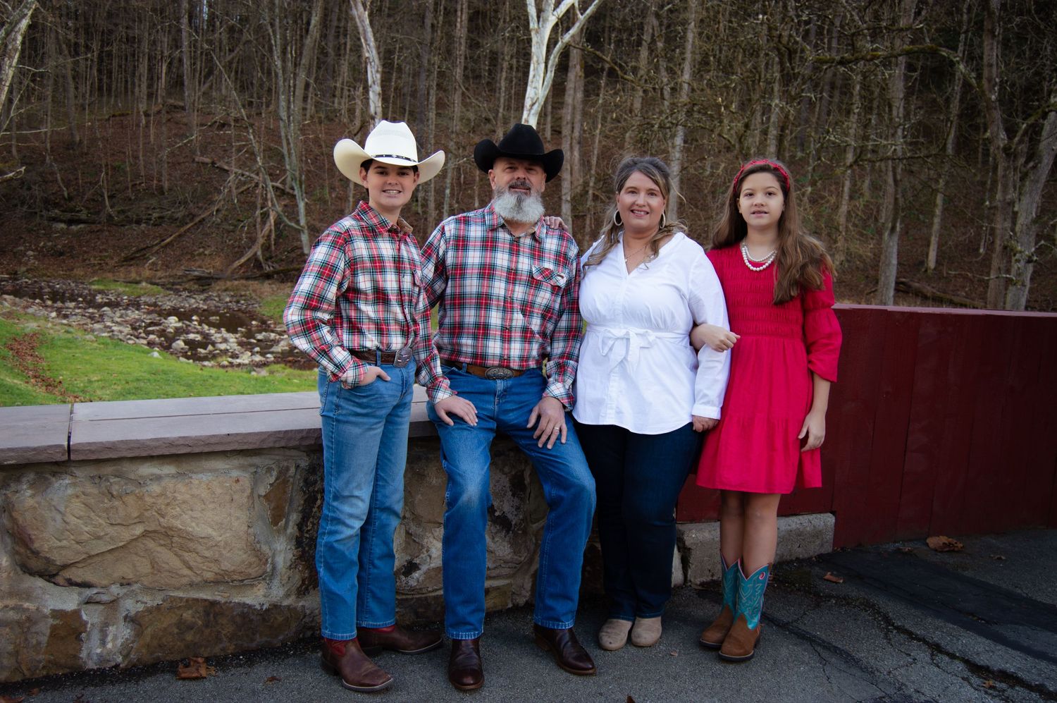Family members wearing casual western attire pose together in front of a rustic red barn in a rural setting.
