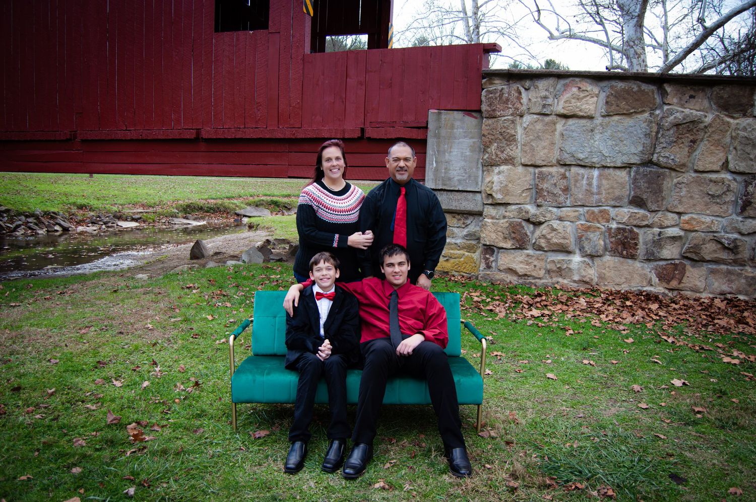 Family members sit together on a teal bench against a rustic stone wall and red barn backdrop.
