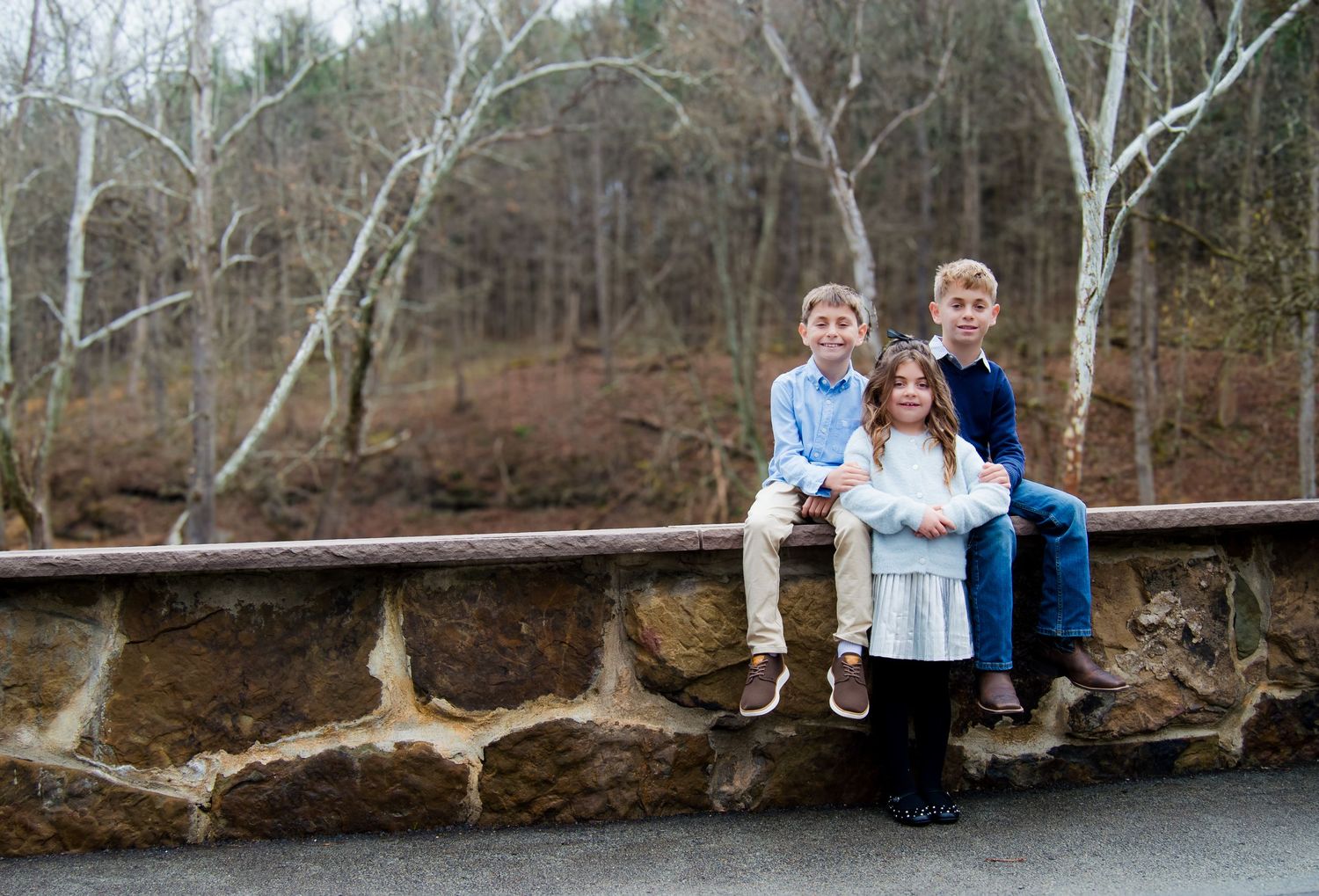 Three children sit together on a stone wall in front of a wooded forest area during winter.