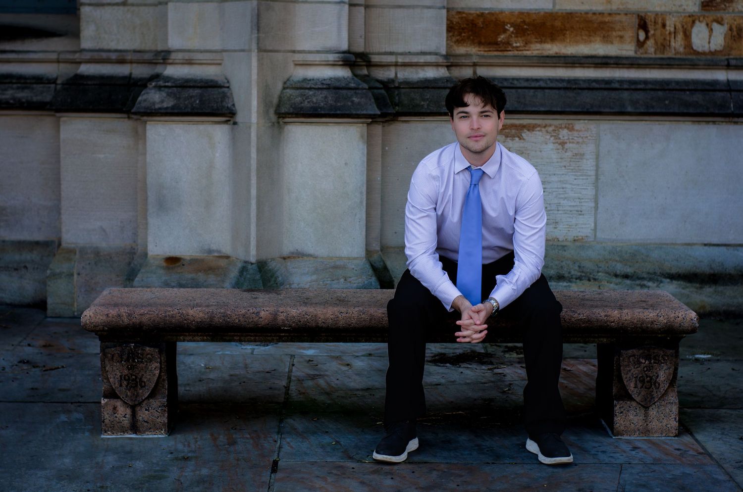 Young man in white shirt and blue tie sitting on a stone bench outdoors near a classical building.