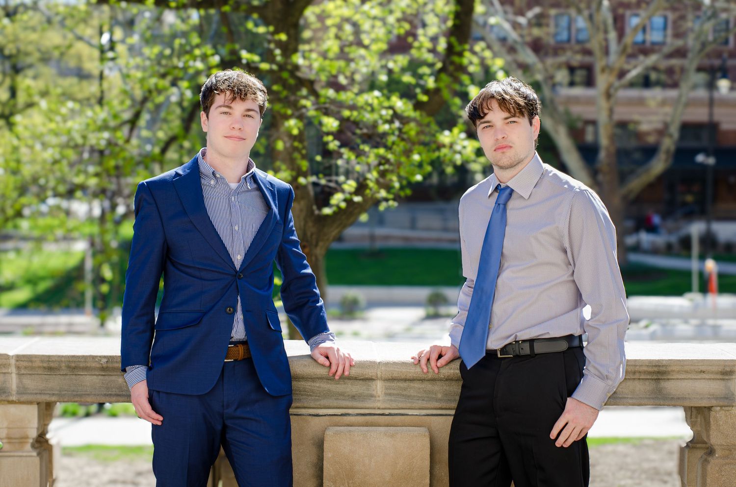 Two young men in formal attire pose outdoors near a stone railing with spring trees in background.
