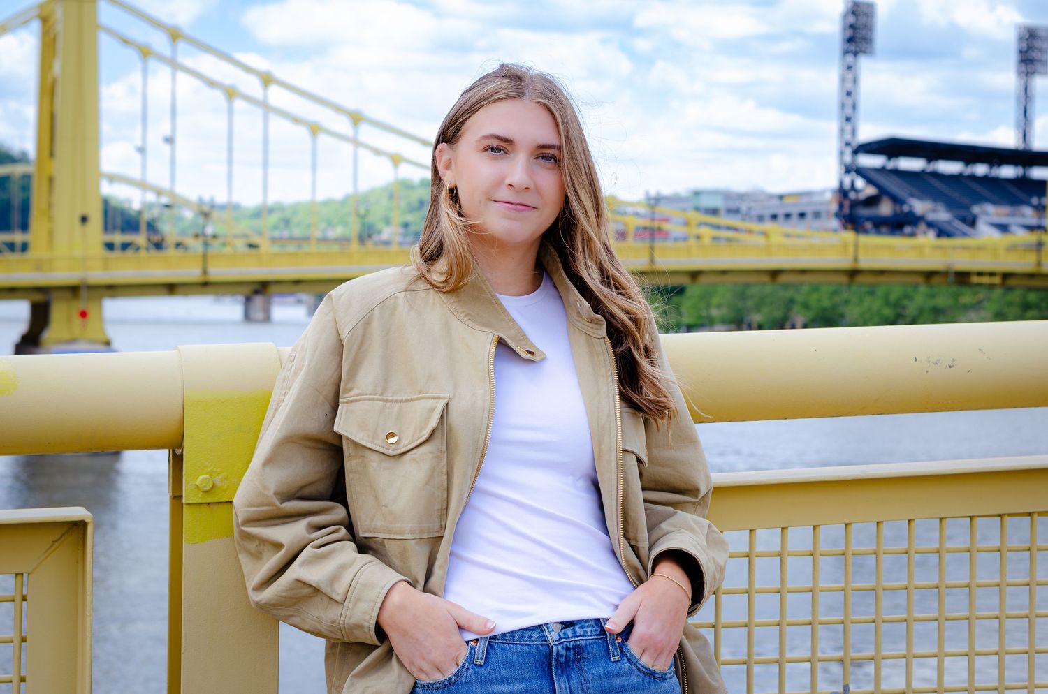 Young woman in tan jacket and jeans leaning on yellow railing of Pittsburgh bridge.