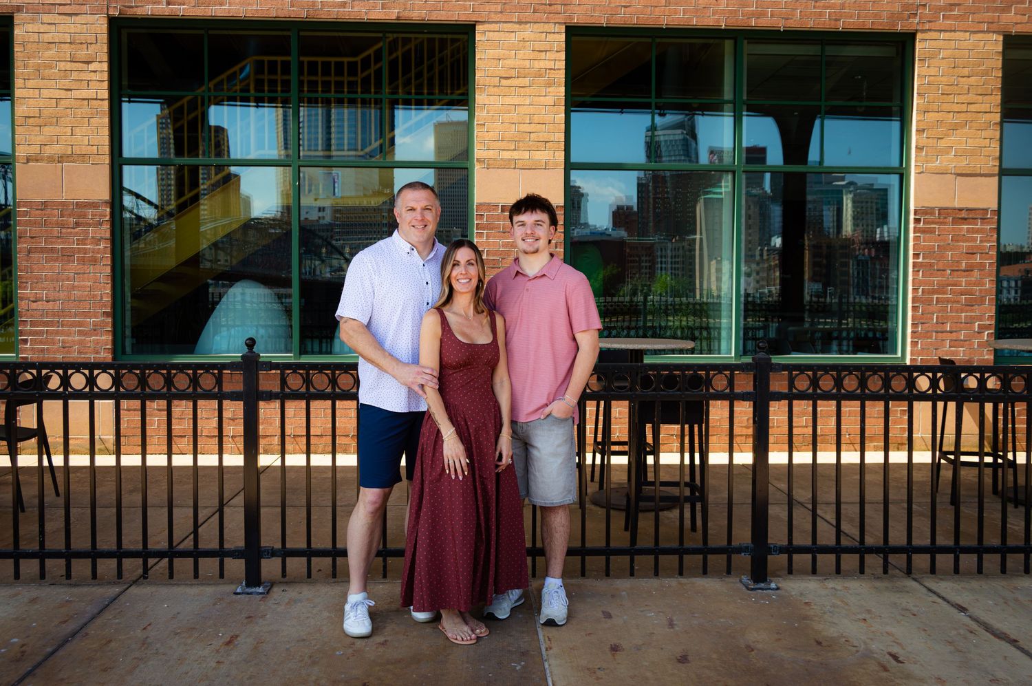 Family of three smiling together outdoors near a brick building with decorative iron fence on a sunny day.