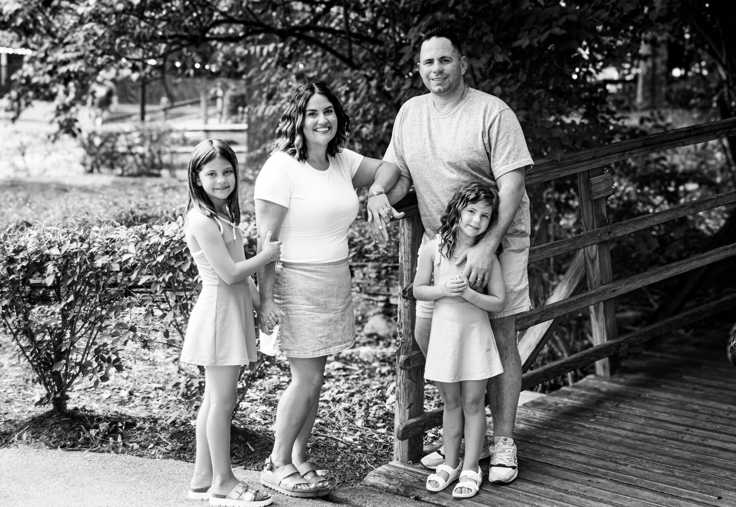 Black and white family portrait of parents with two young daughters posing on a wooden bridge surrounded by lush greenery.