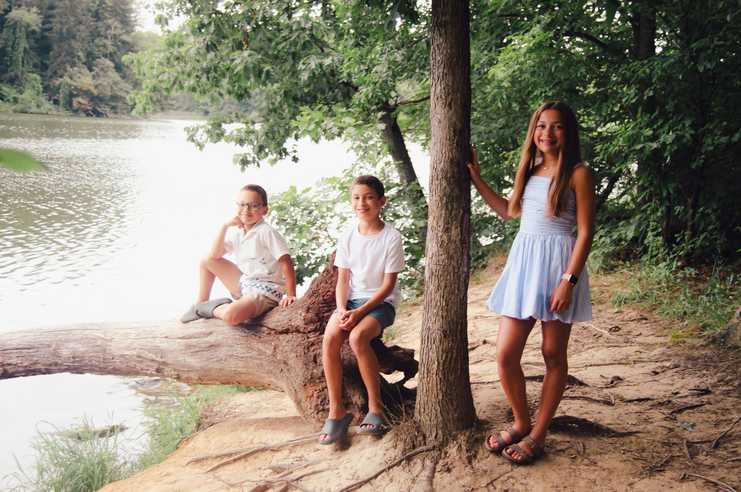 Three people enjoying a summer day by a peaceful lake, sitting and standing near a tree on the shoreline.