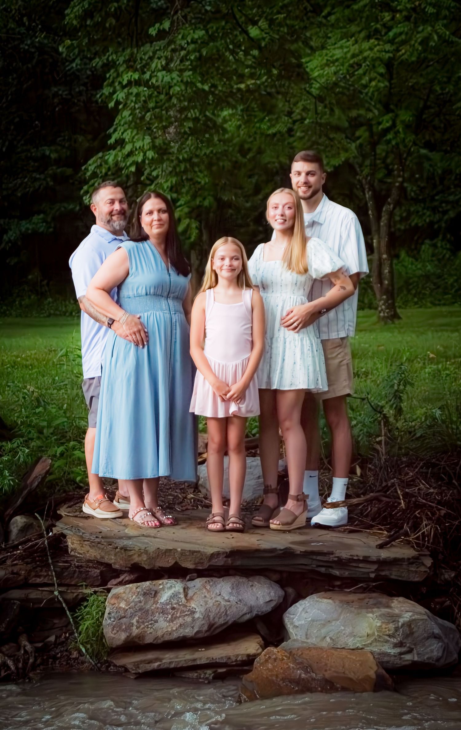 A family in coordinated pastel outfits stands together on large rocks in a wooded outdoor setting.
