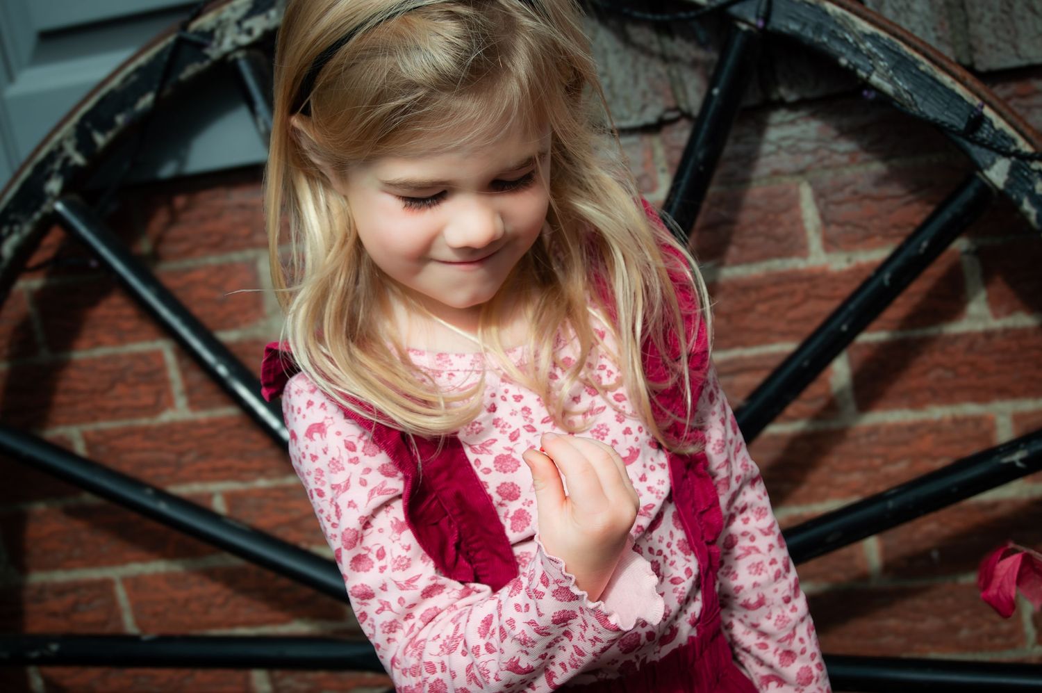 A young child in a pink floral top and burgundy overalls stands shyly against a brick wall with a wagon wheel.